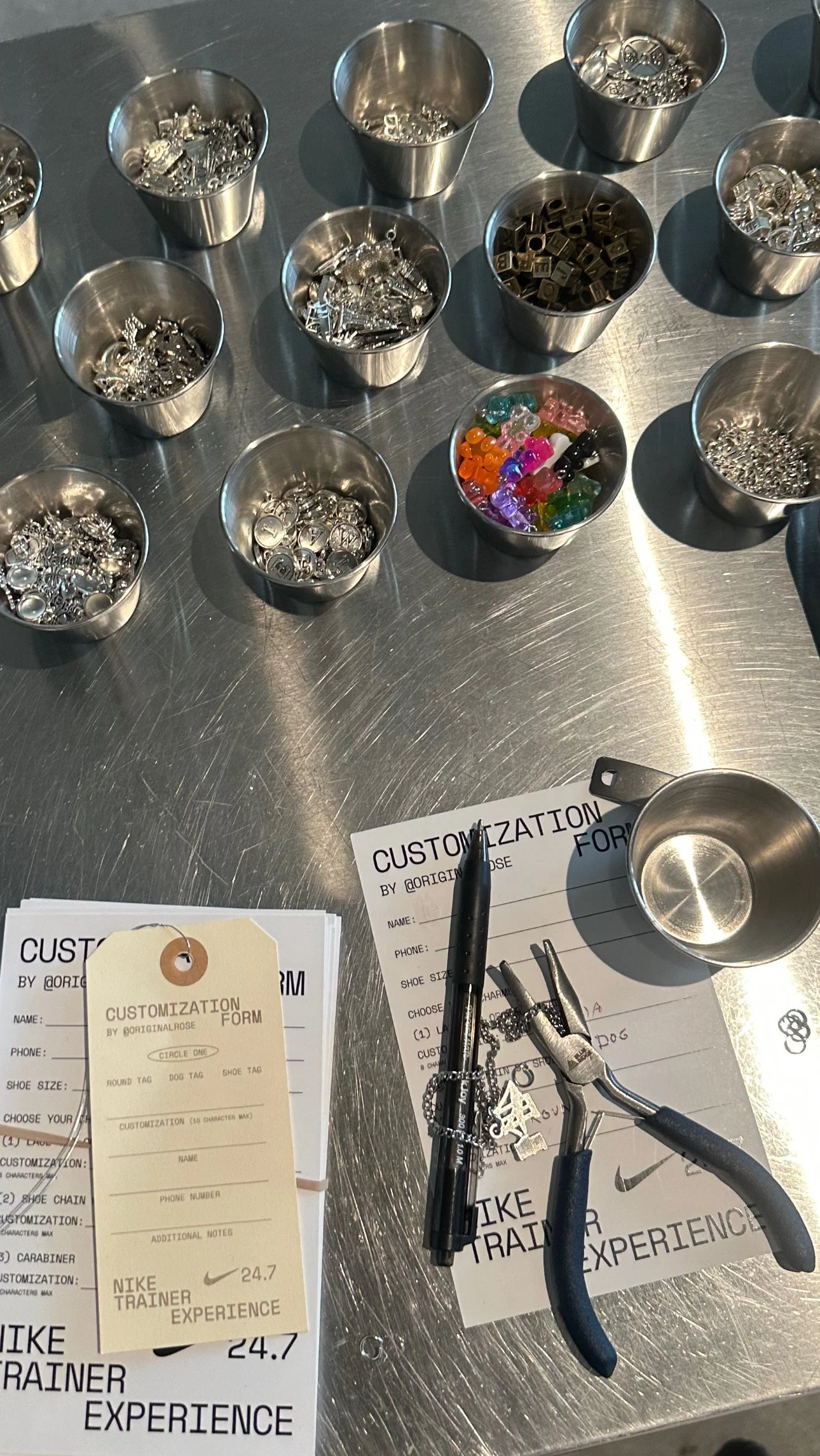 A metal table with small tins containing various jewelry charms and decorative items, a colorful assortment of glass beads, jewelry-making tools, and sheets of customization forms for personalized jewelry design.