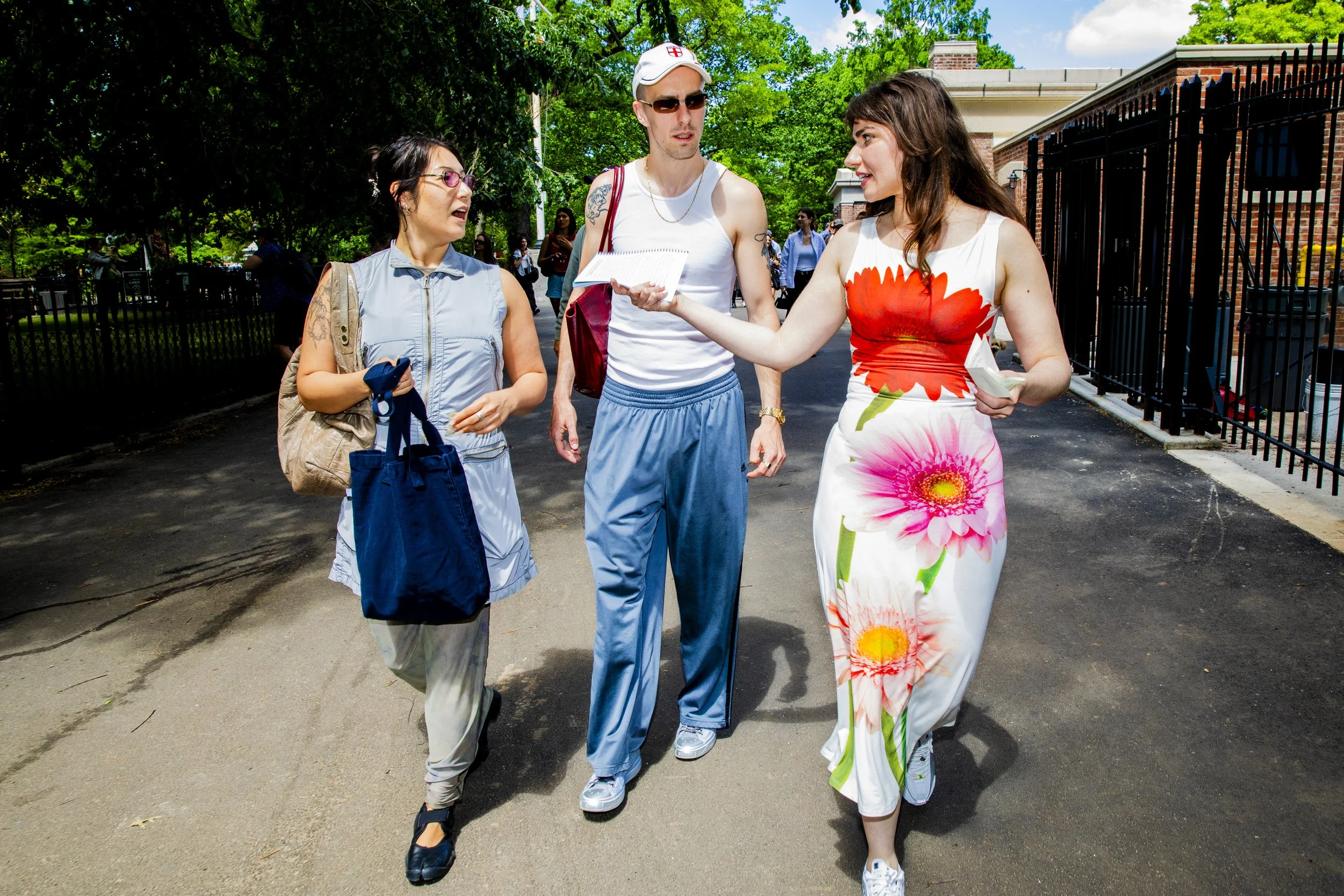 Three people walking outdoors on a sunny day, two women and one man, engaged in conversation, with green trees and buildings in the background.