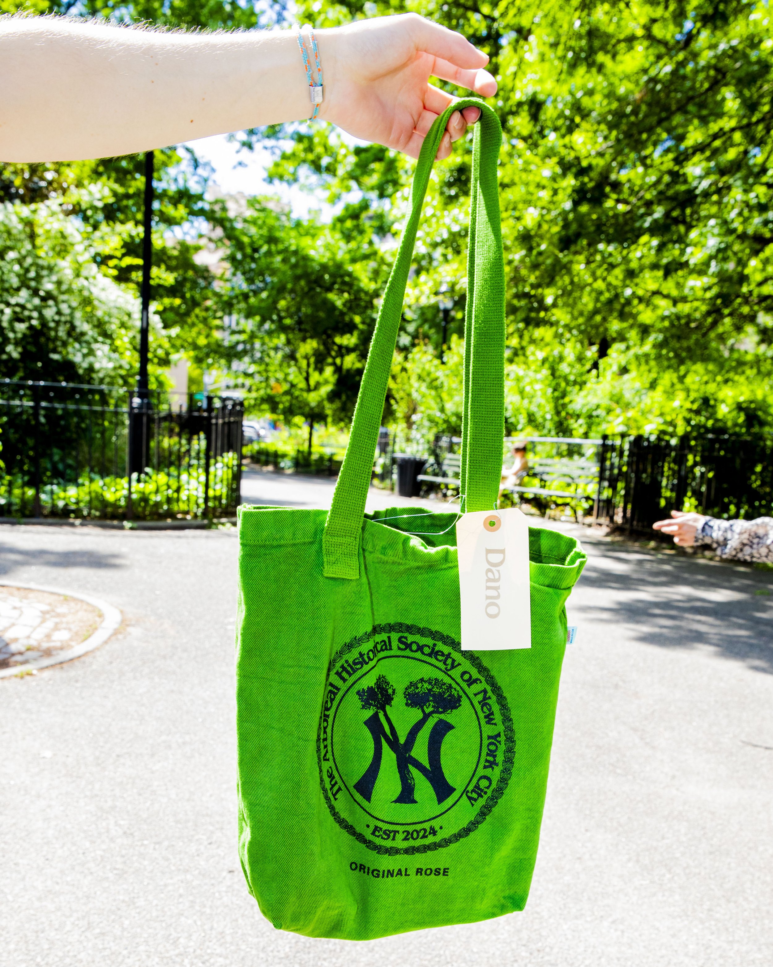 A person's hand holding a bright green tote bag with black logo and text, set outdoors on a sunny day with trees and a park pathway in the background.
