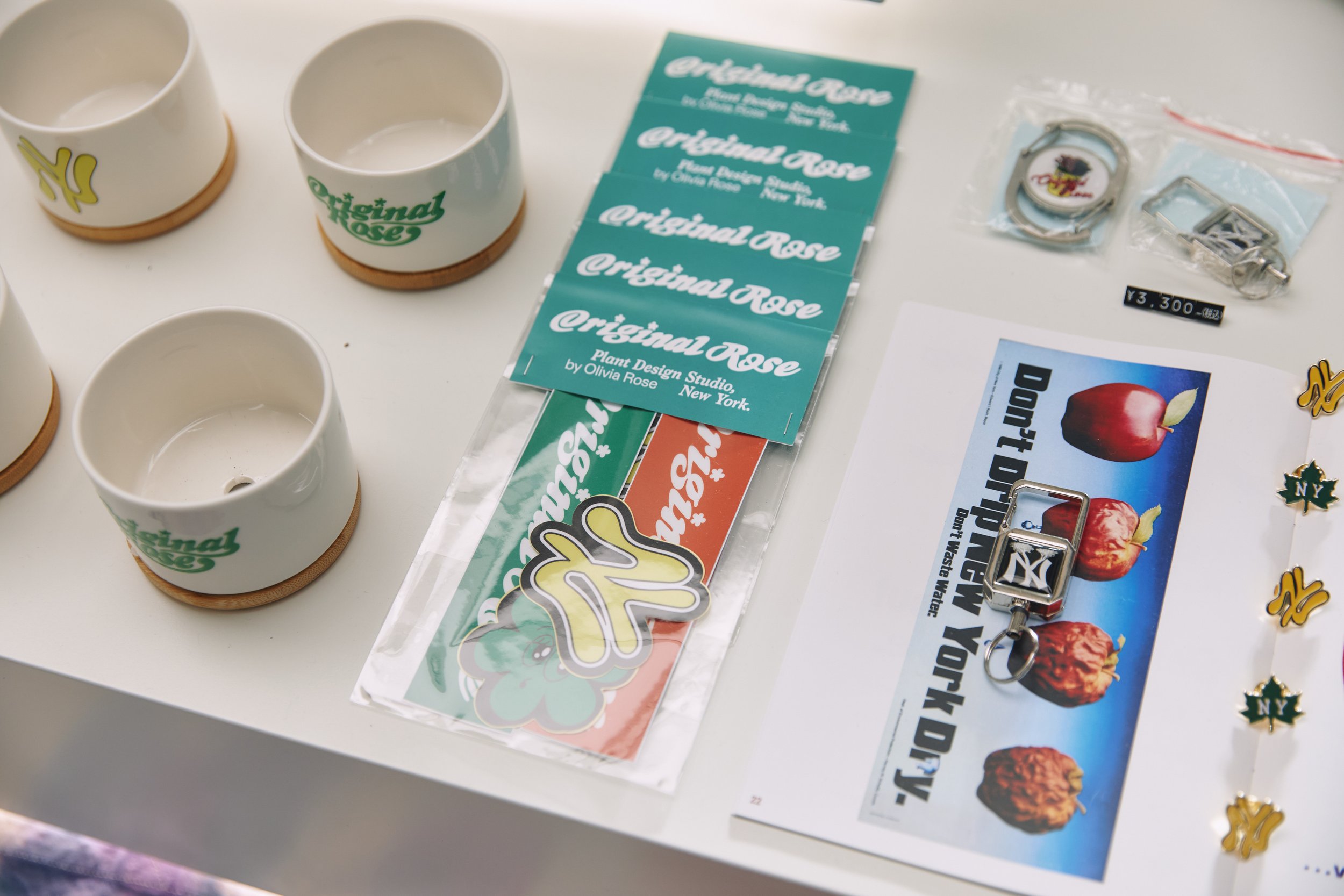 Set of ceramic cups with the 'Original Rose' logo, plant stickers, a watch, and a card with apples and partnered snacks on a white surface.