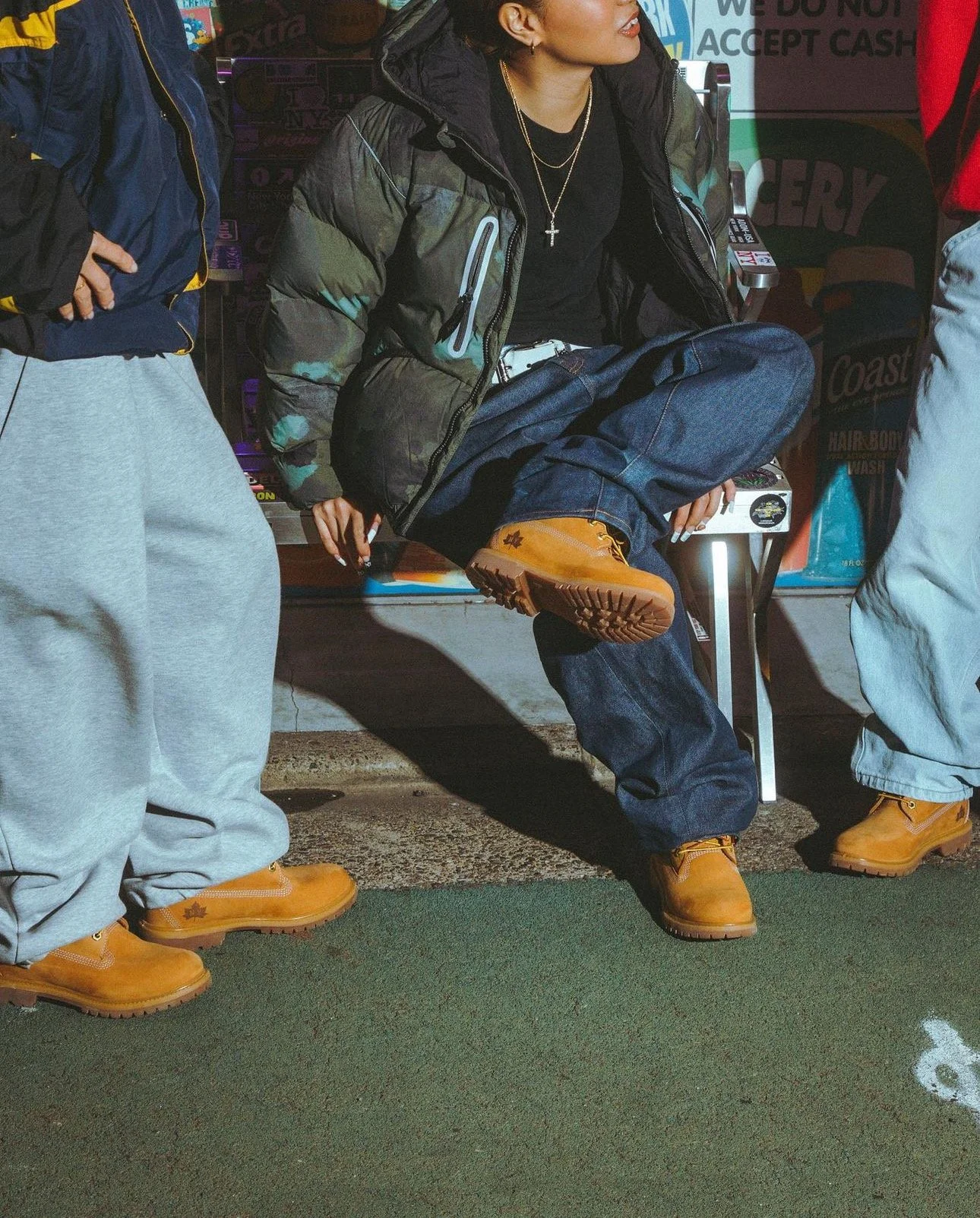 Person sitting on a bench wearing a black jacket, gold boots, and jewelry, surrounded by two other people, in front of a storefront with signs about accepting cash and other products.