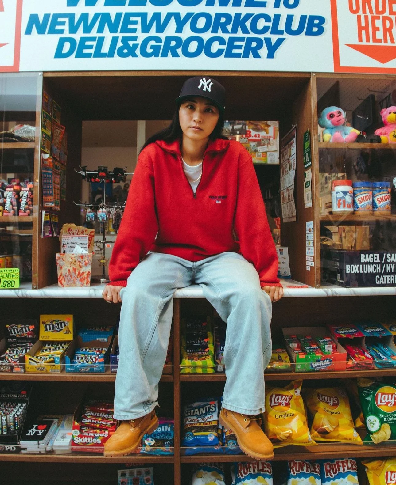 A woman sitting on a counter in a convenience store, wearing a black New York Yankees cap, red fleece jacket, and tan boots, surrounded by snacks and candy.