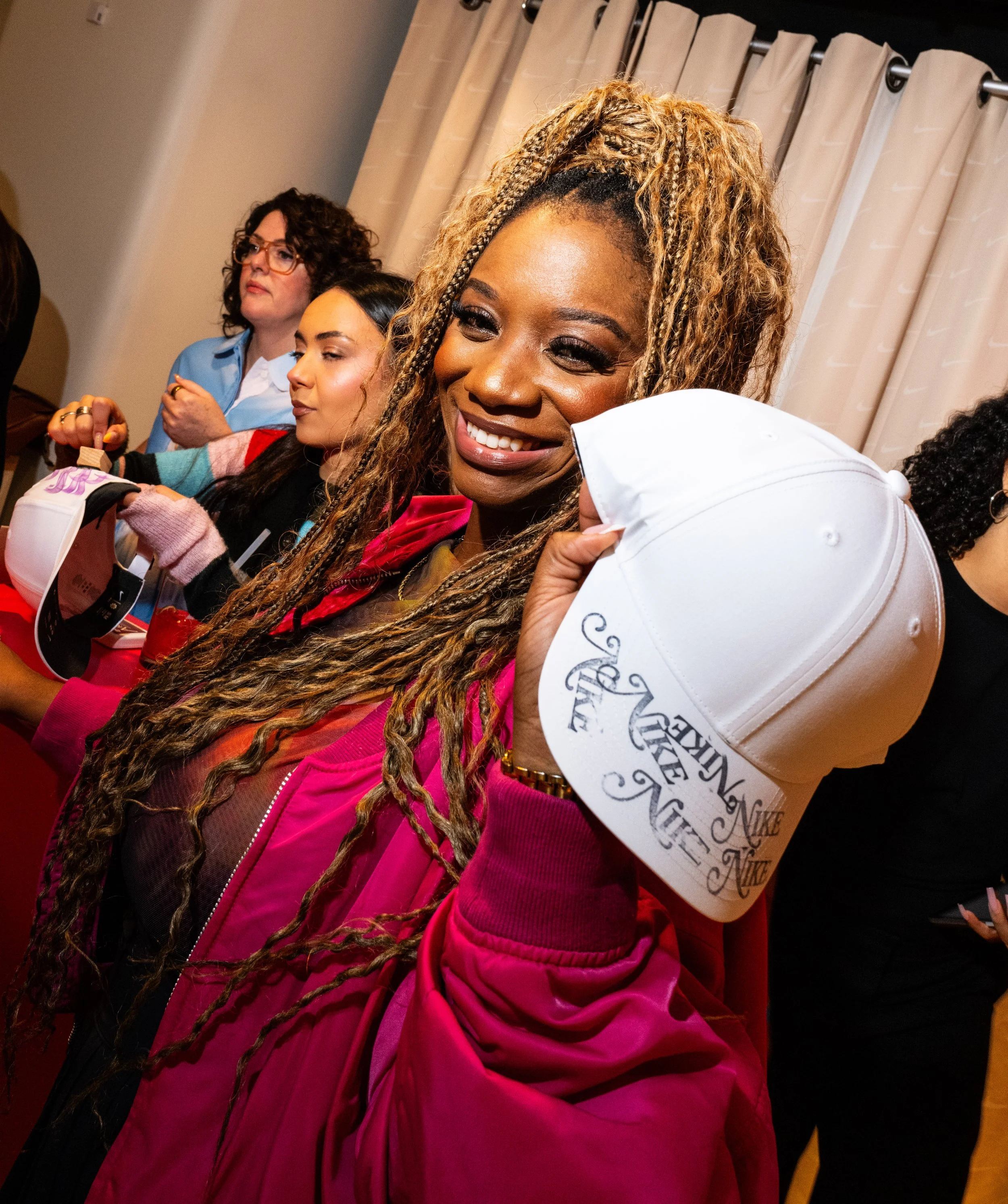 A woman smiling and holding a white Nike cap, surrounded by other women at an indoor event.