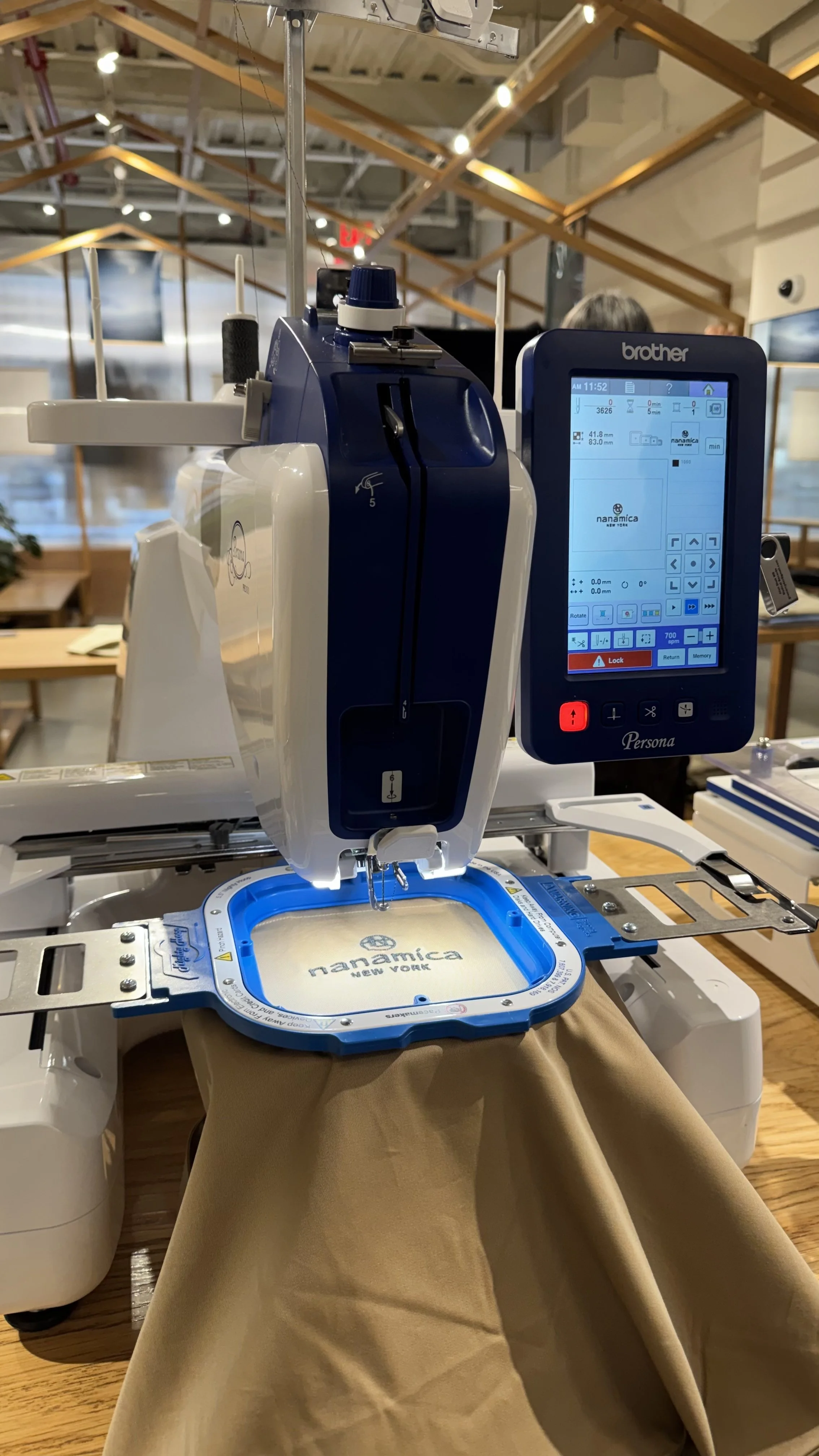 A Brother sewing machine with a digital display, embroidering a fabric piece with "namamica NEW YORK" on it in a well-lit indoor setting.