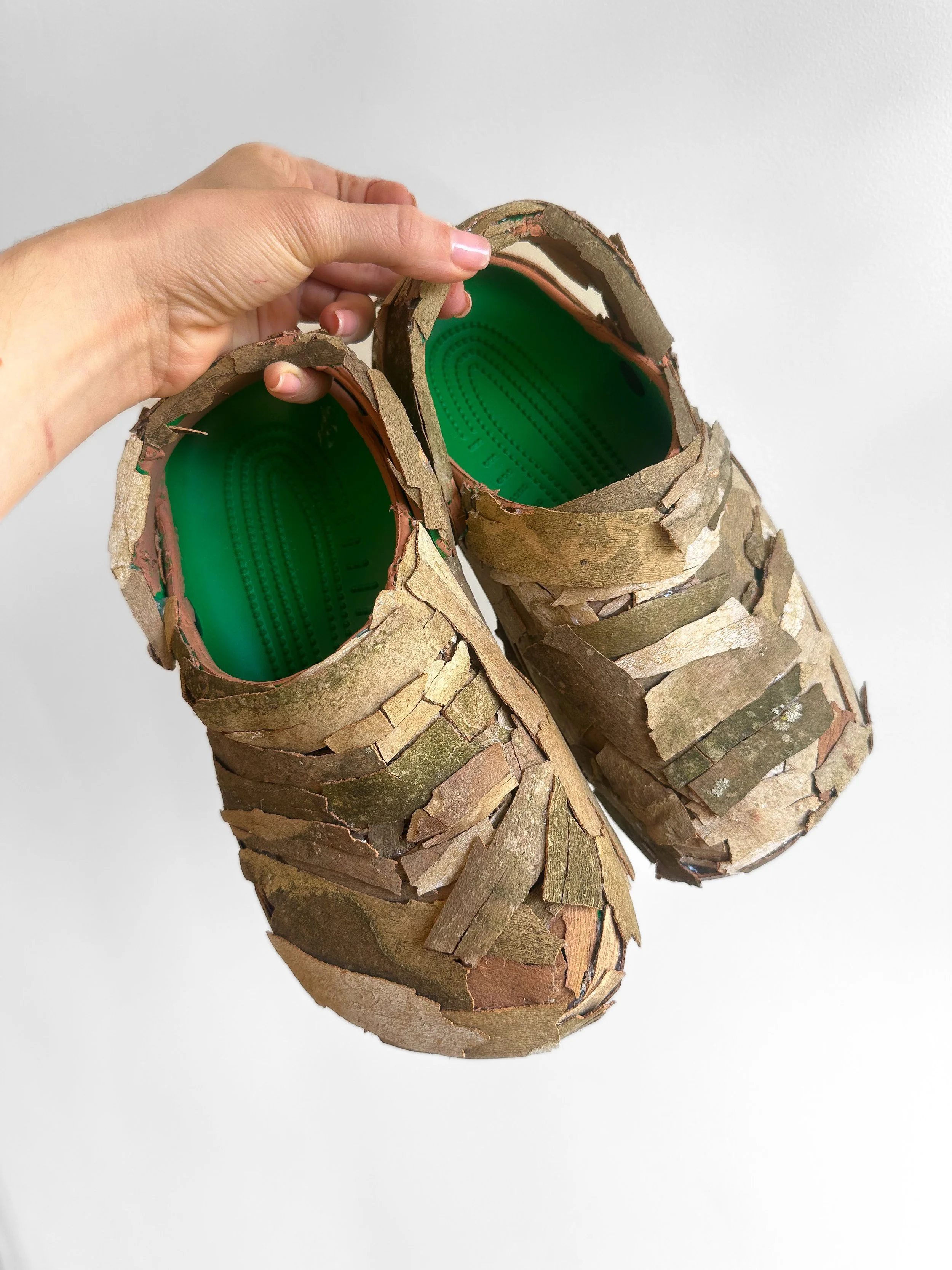 Pair of shoes covered in irregularly arranged pieces of bark with green insoles, held in a person's hand against a plain white background.