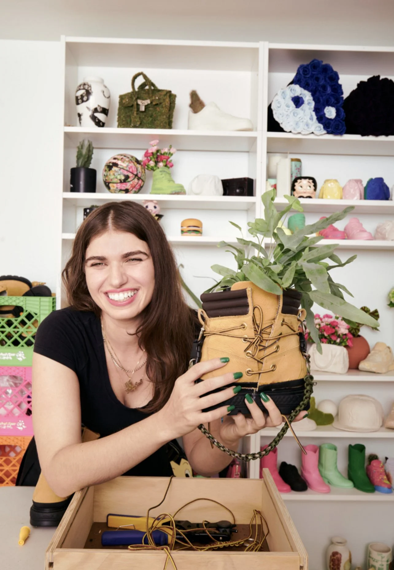 A smiling woman with long brown hair holding a tan and black hiking boot with a healthy green plant inside. She is in a colorful crafting or vintage store with shelves of decorative items and shoes in the background.