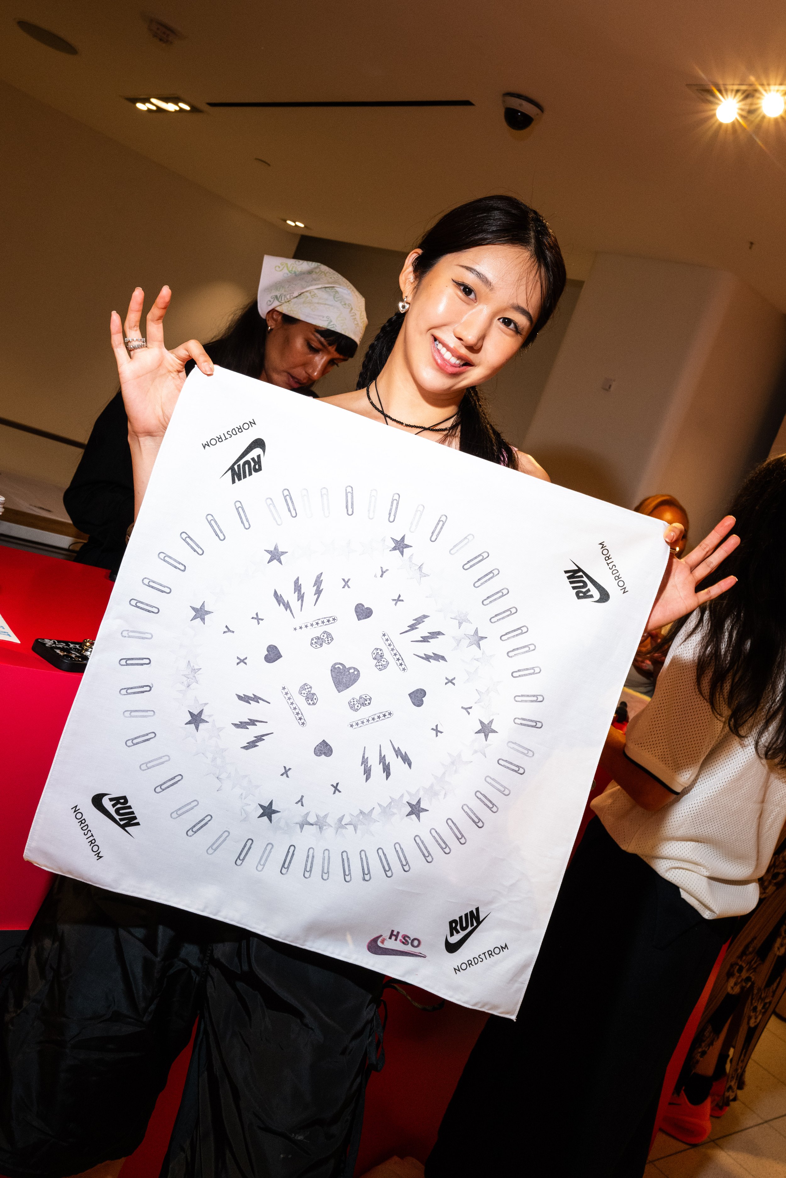 A woman holding a large white bandana with various printed designs and logos, smiling and making an OK gesture with her hand at an indoor event.