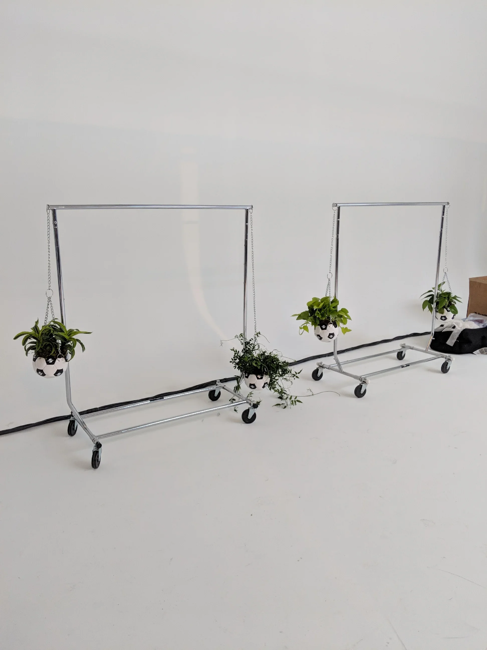 Two metal clothing racks on wheels, each with a potted plant in a soccer ball decorated planter hanging from the top bar, with a white wall background.