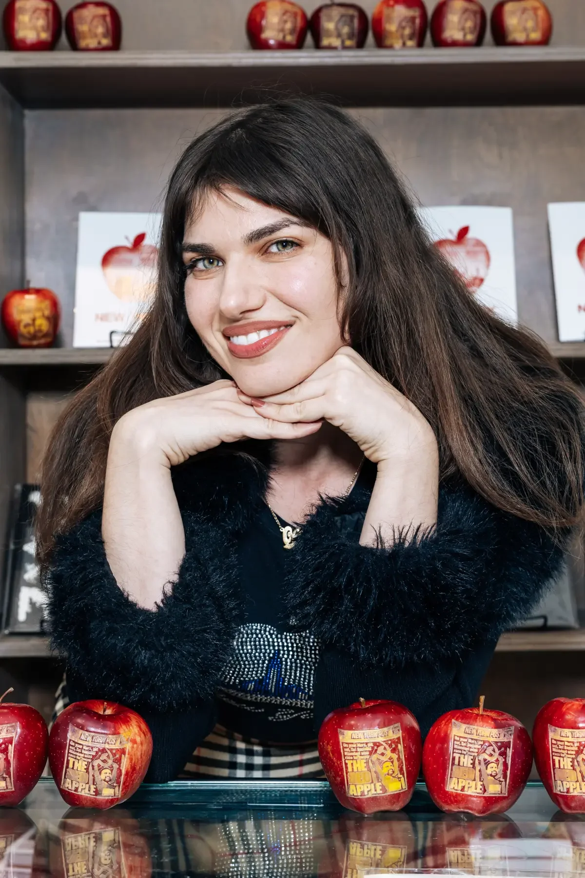 A woman with long dark hair, smiling, resting her chin on her hands, sitting behind a row of red apples on a table, with a background of shelves with red apple-themed decorations and signs.
