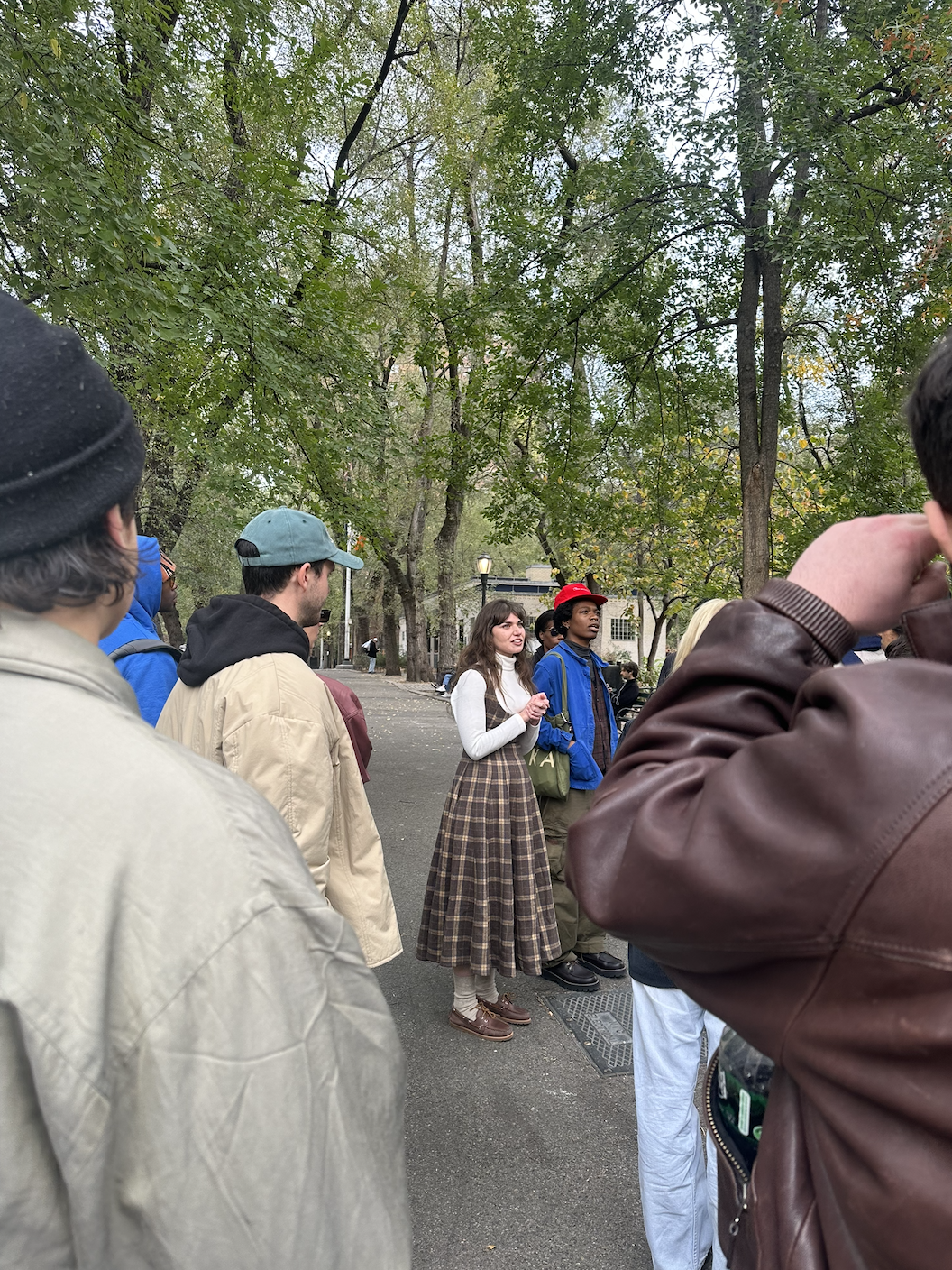 Group of people outdoors in a park, listening to a woman speak. The scene features trees with green leaves, and the group appears diverse in age and appearance.