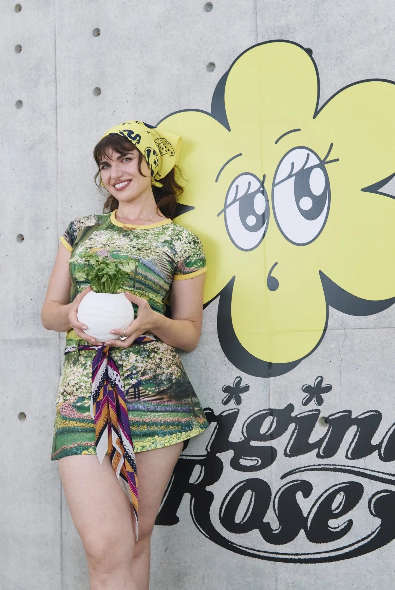 A woman with dark hair wearing a yellow headscarf and a colorful dress holding a potted green plant, standing next to a large cartoon flower on a gray wall.