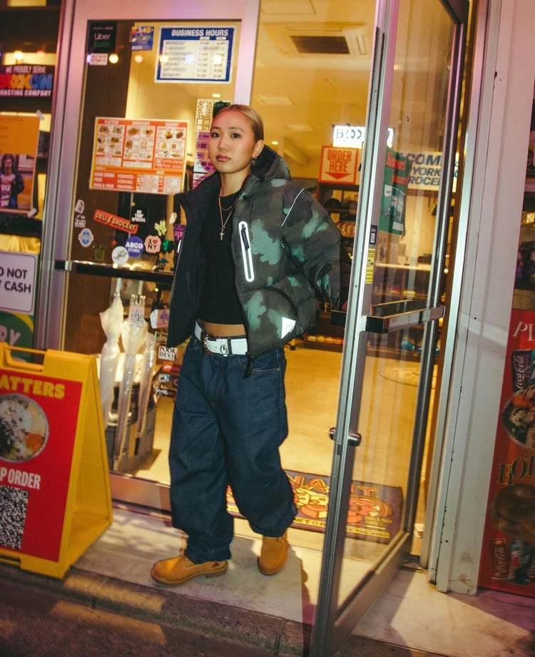 Model in a camouflage jacket and yellow boots outside a convenience store