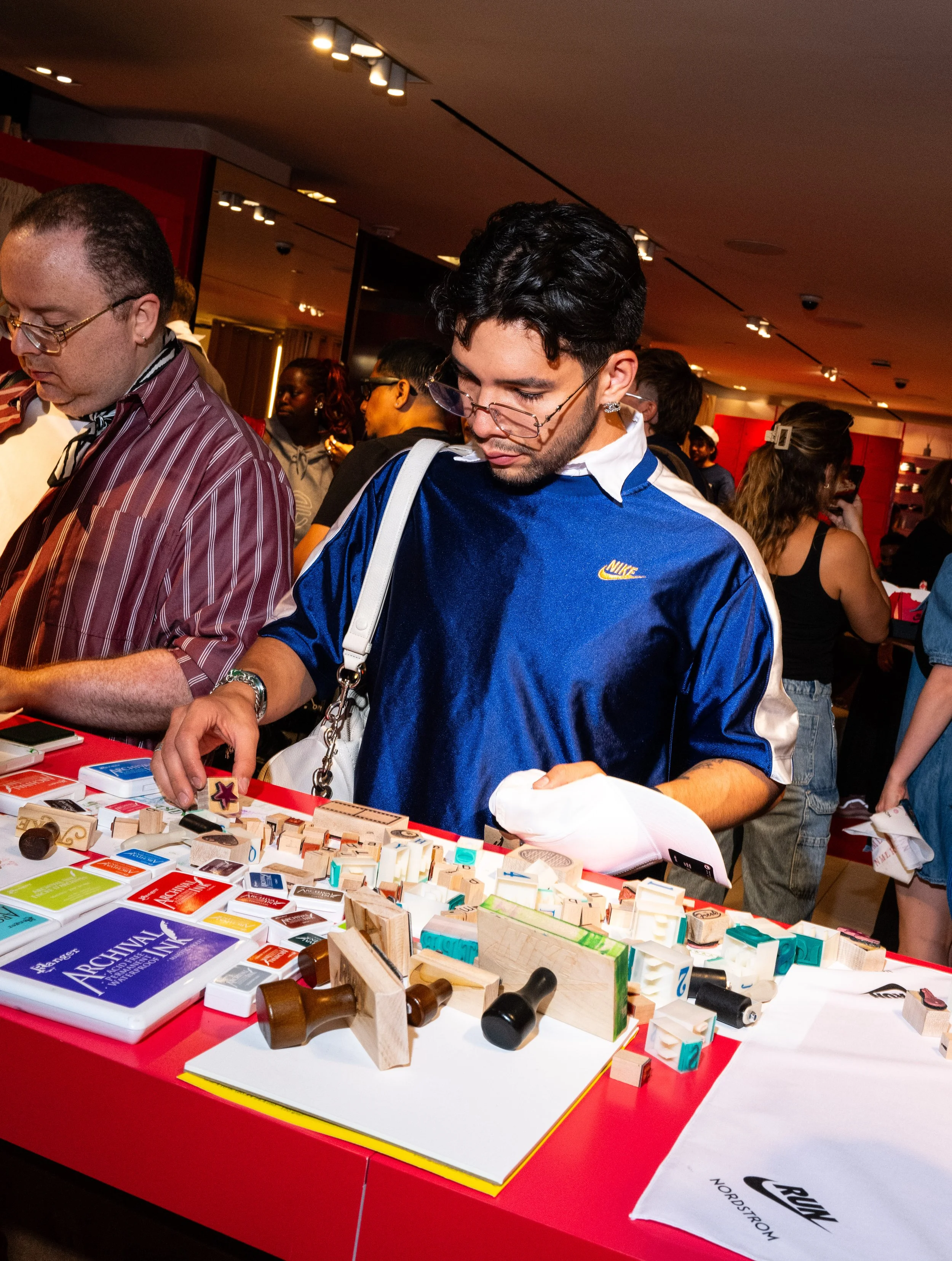 A young man with glasses, a blue Nike sports shirt, and tattoos on his arm, shopping at a market stall with rubber stamps and ink pads, surrounded by other shoppers in a busy indoor setting.