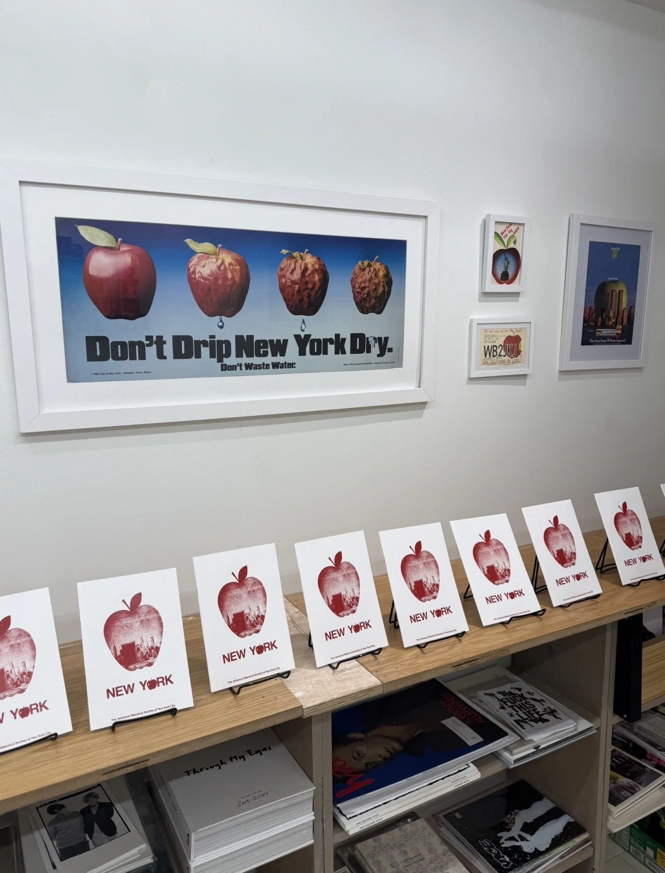 Gallery wall with four framed art pieces, the largest displaying rotting apples labeled 'Don't Drip New York Dry.' Below is a display of postcards or flyers with a red apple and New York City skyline, on a wooden table.