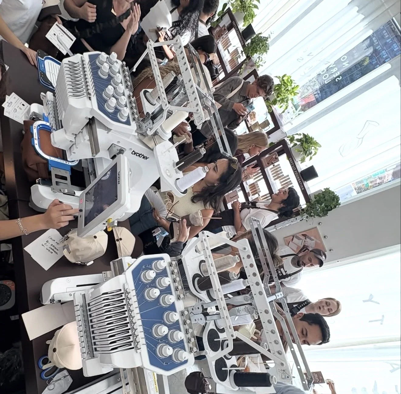 People in a classroom observing and working with Brother industrial sewing machines, with some individuals taking notes and others looking at the machines, near large windows and urban scenery outside.
