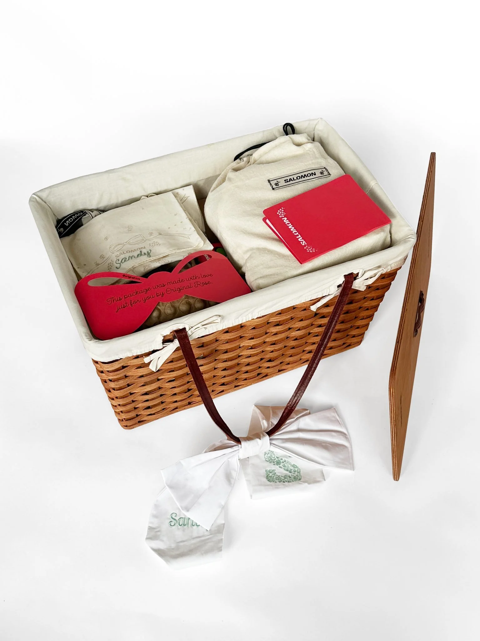 A woven basket containing various packaged gift items, including cloth bags, a red heart-shaped card, and a white cloth bag, with a white ribbon tied around the handle and a wooden panel leaning against the basket.
