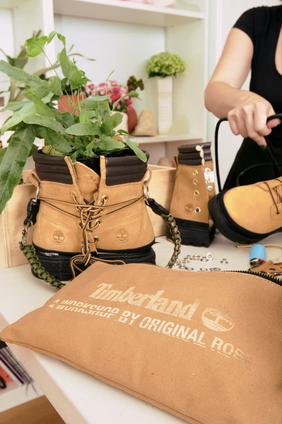 A pair of Timberland boots used as planters with green plants inside. The boots are on a table with various tools and accessories nearby, and a tan Timberland branded bag in the foreground.