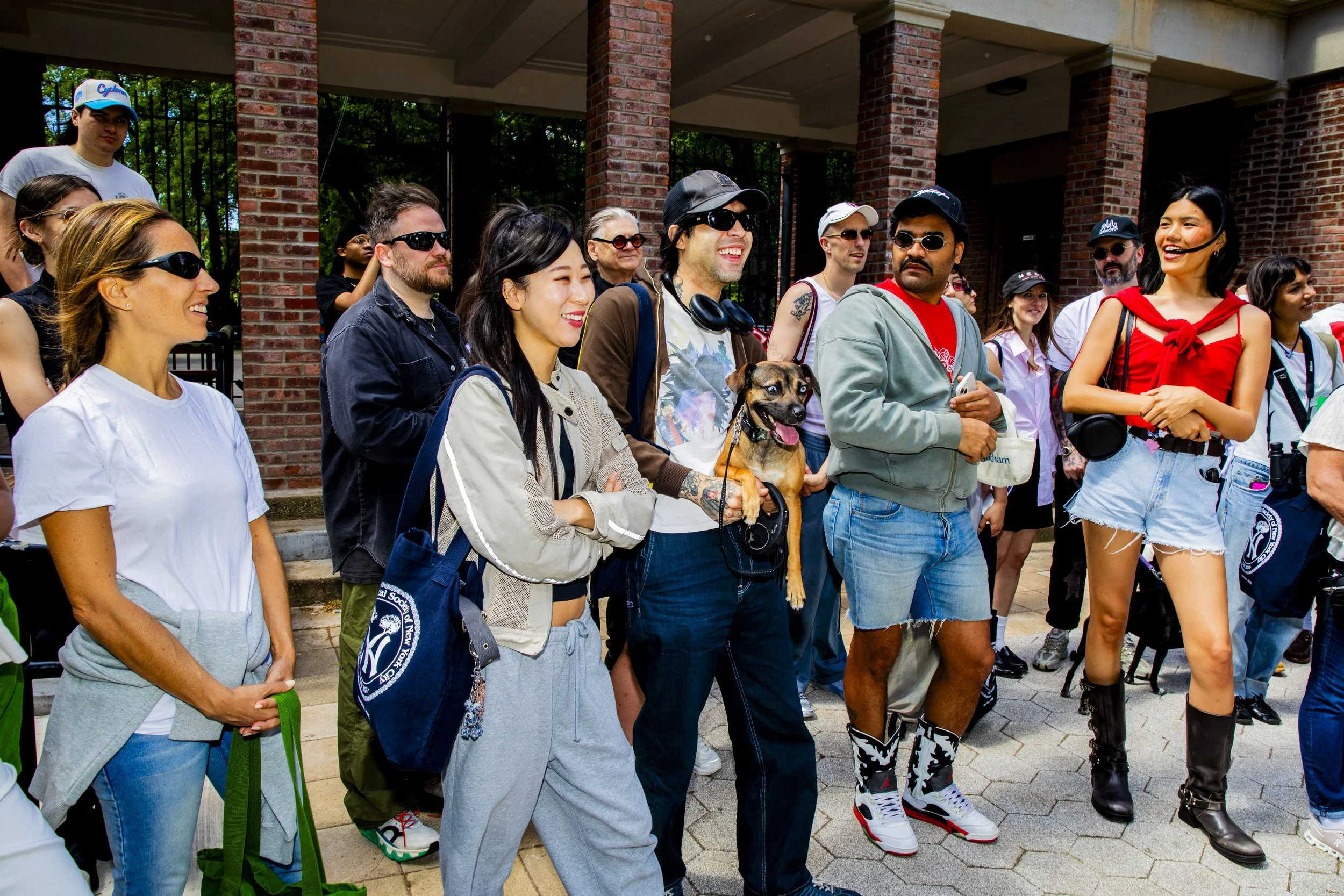 Group of diverse people gathered outdoors, smiling and engaged, with some wearing sunglasses, casual clothing, and carrying backpacks. A woman holds a dog, and others are standing under a brick structure.