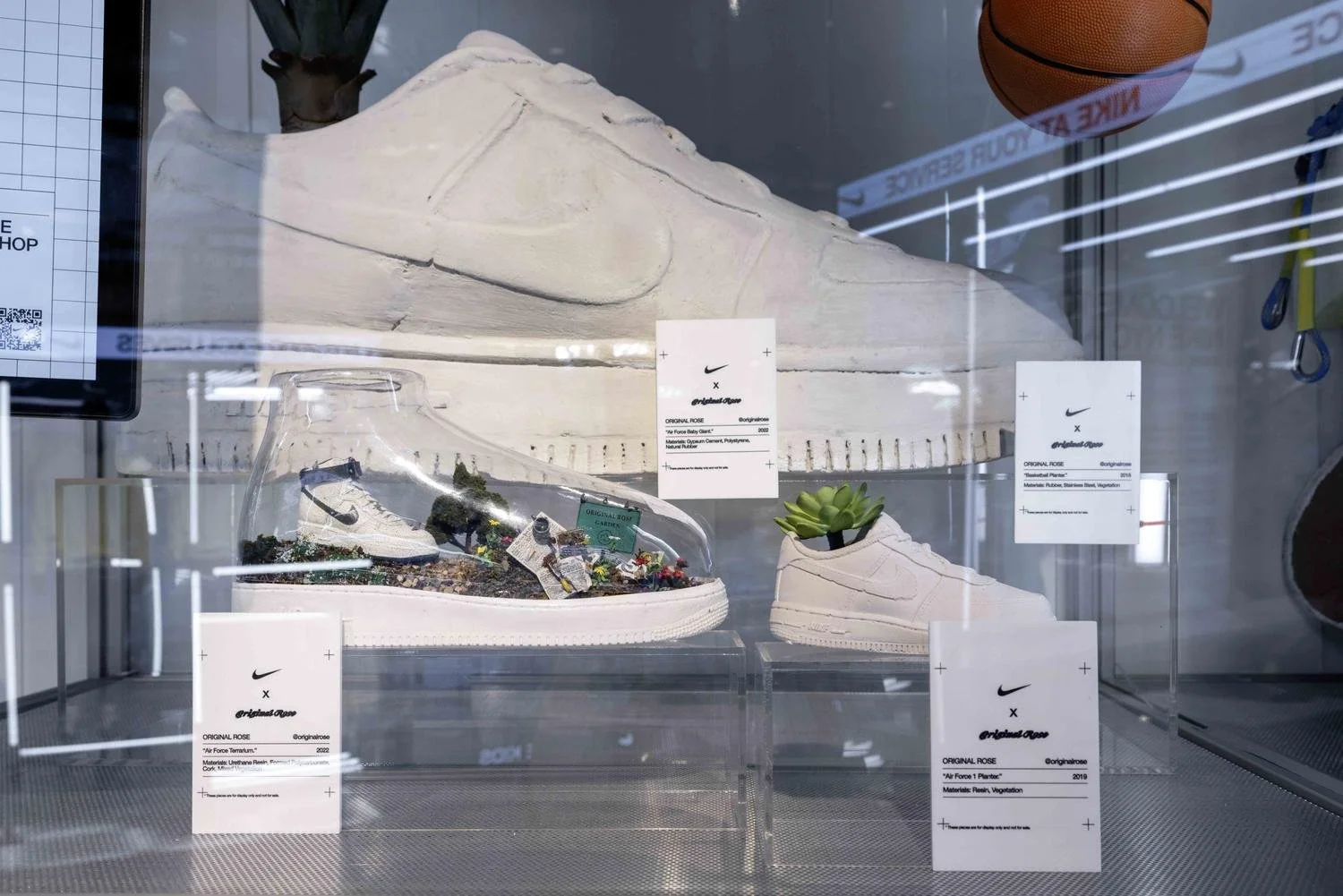 Display case featuring a Nike sneaker and a sneaker with miniature landscape and plant decor, labels with product details, and a large white sneaker in the background.