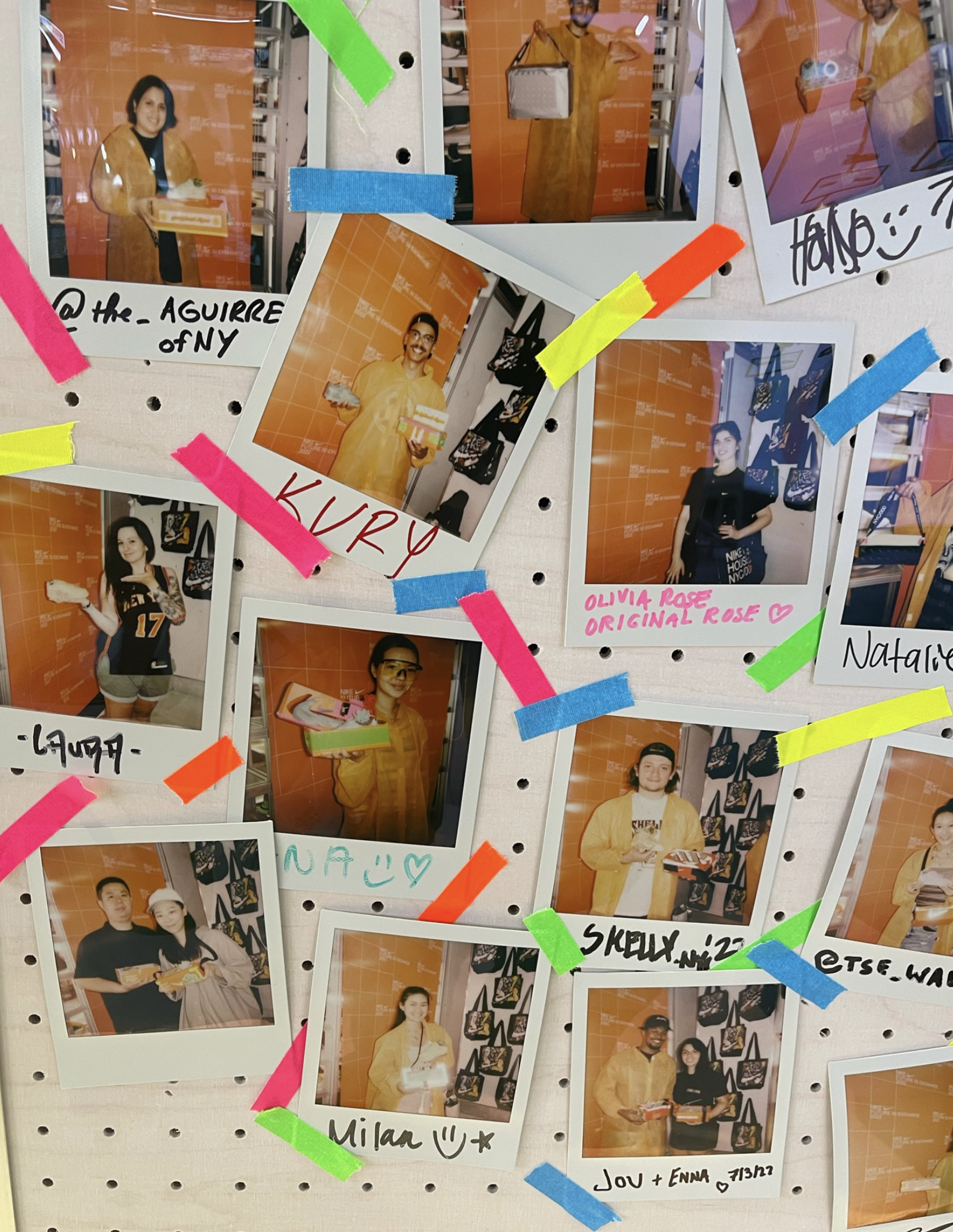 A collage of Polaroid photos placed on a white pegboard, with each photo showing different people holding boxes, some smiling. The photos are attached with colorful tape and have handwritten names and messages on them, set against an orange backgroun