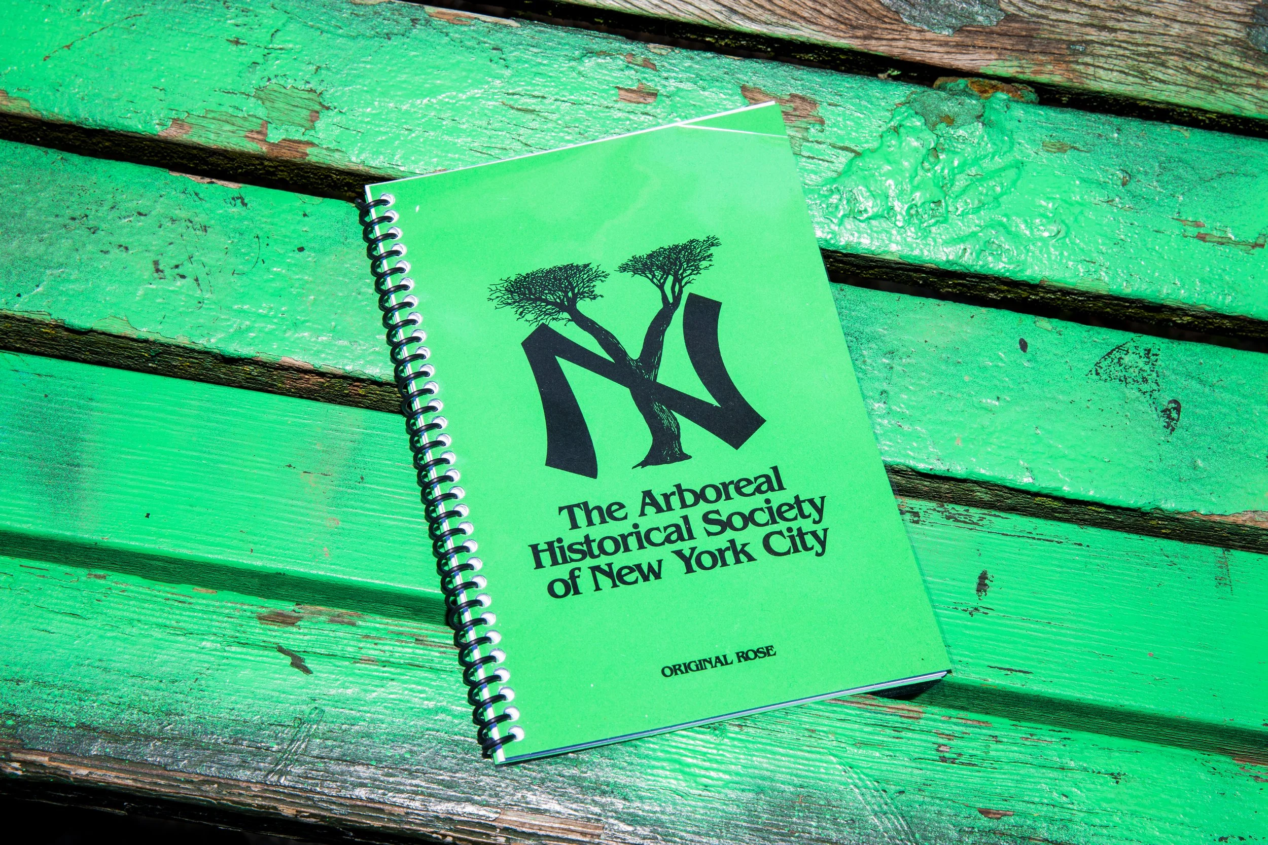 Green spiral notebook with a logo of trees and text reading 'The Arboreal Historical Society of New York City' on a weathered green wooden surface.