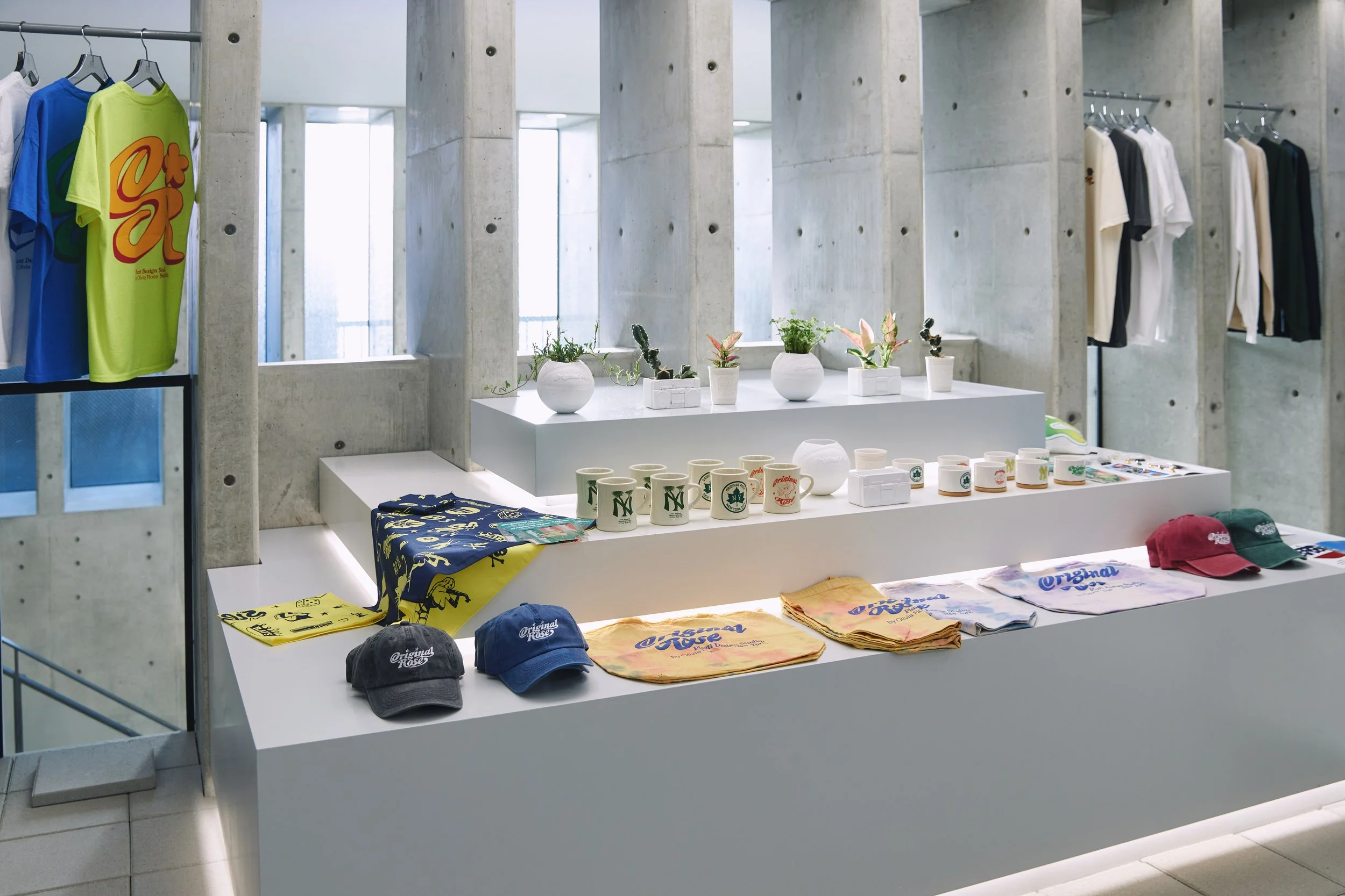 Display of souvenirs in a retail store, featuring graphics on T-shirts, caps, and mugs, with potted plants on a white table.