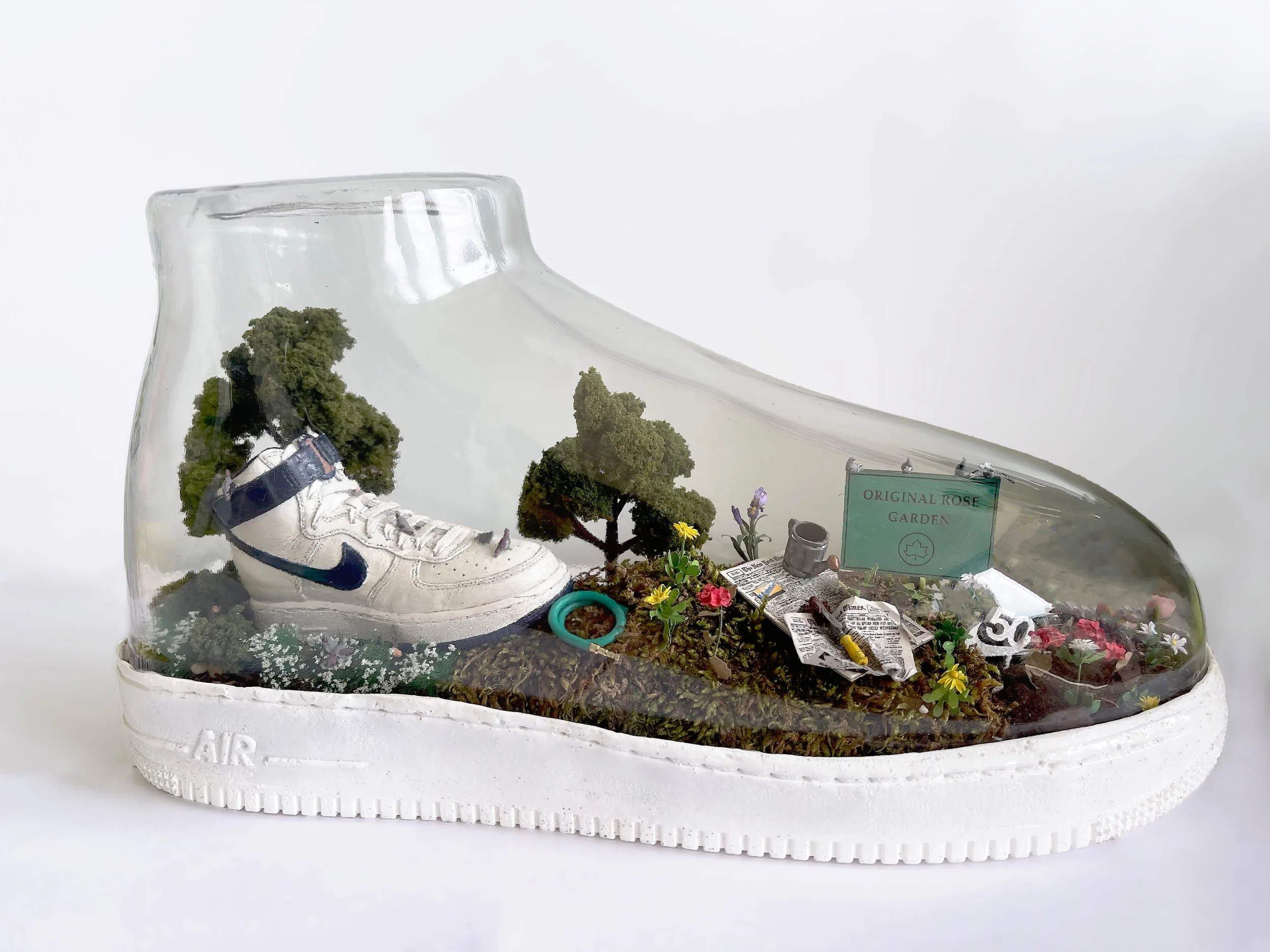 Miniature landscape inside a clear Nike sneaker, featuring small trees, flowers, a newspaper, a watering can, and a sign that reads 'Original Rose Garden'.