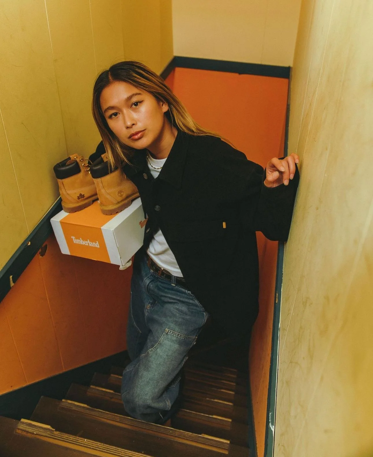 Young woman with light brown hair posing on a staircase, holding a Timberland shoebox with a pair of Timberland boots on top, in a colorful, enclosed space.