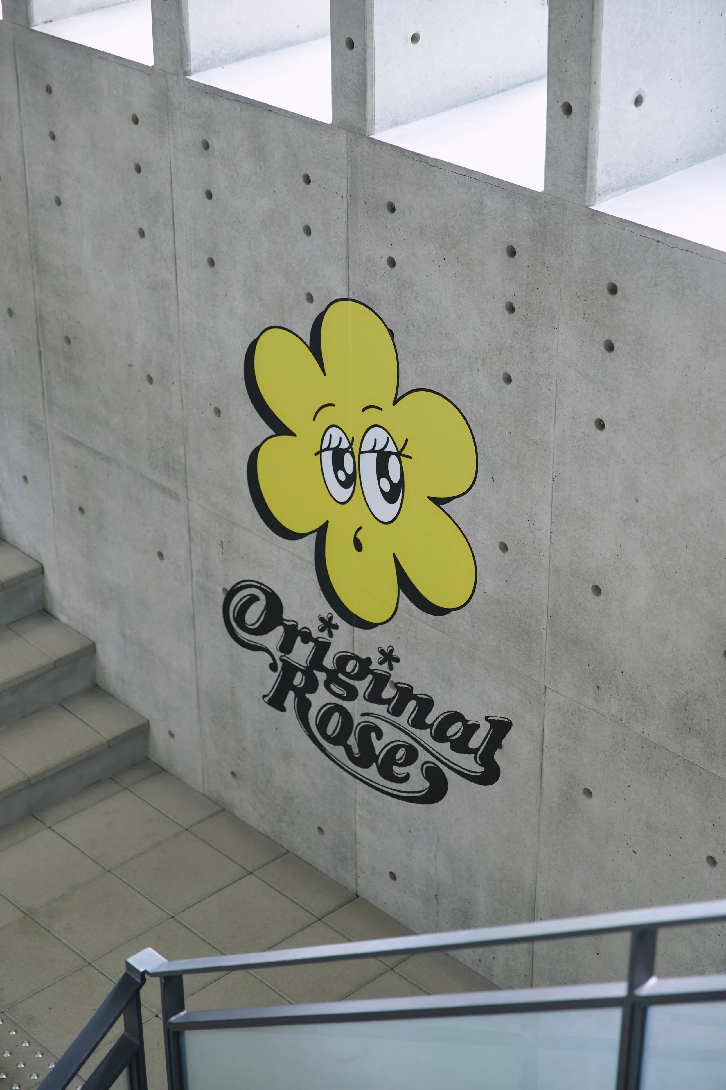 A yellow cartoon flower with big eyes and a small nose on a gray concrete wall. Below it, the text reads ' Annual Rose' with stylized font.
