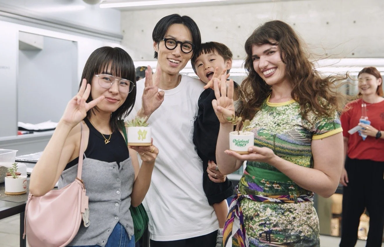 Group of four people smiling and posing at an indoor event, holding small potted plants, making peace signs, with a fourth person in the background.