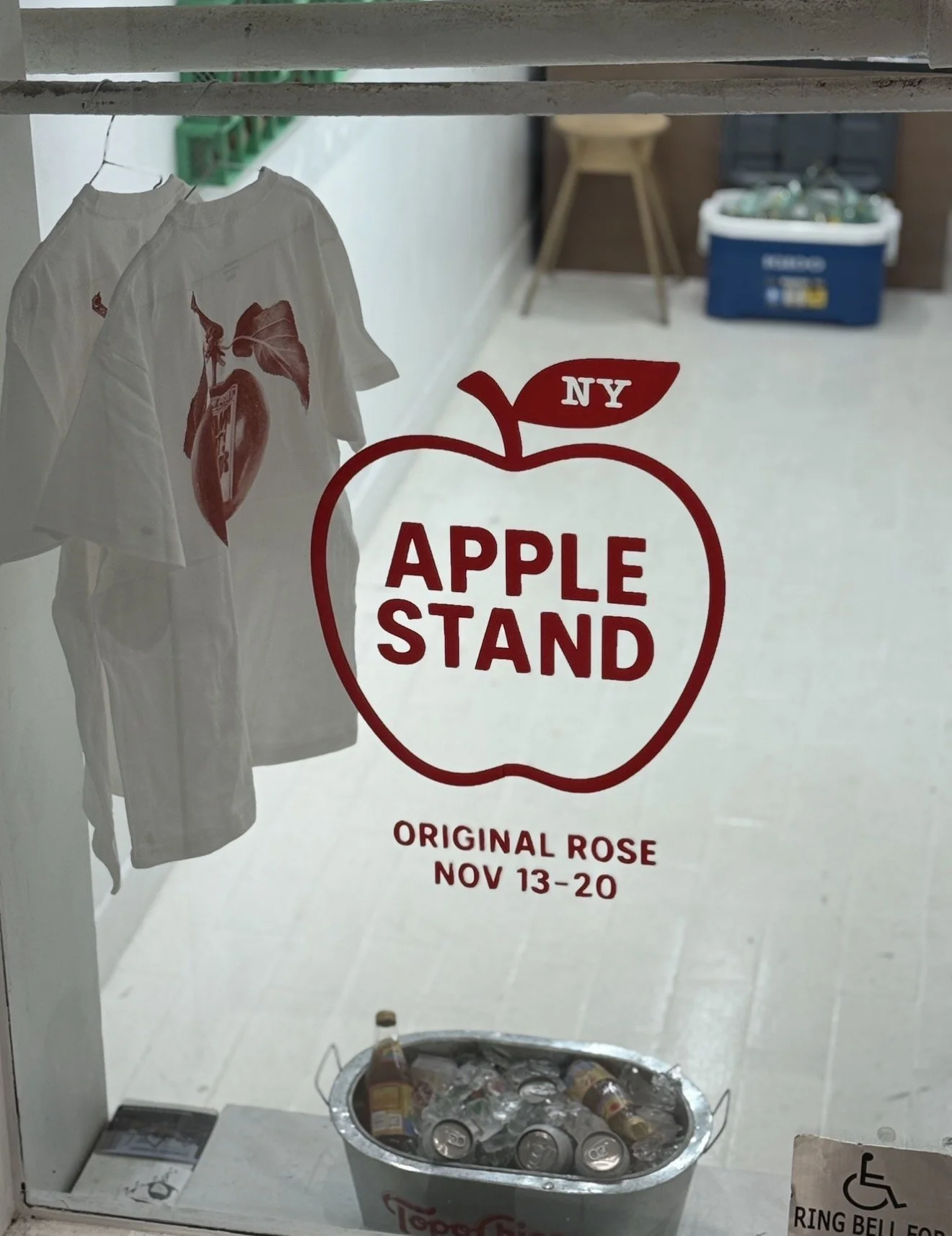 Storefront window for Apple Stand, promoting original roses from November 13 to 20, with white T-shirts featuring a rose design hanging inside and a bucket of bottled beverages on the counter.