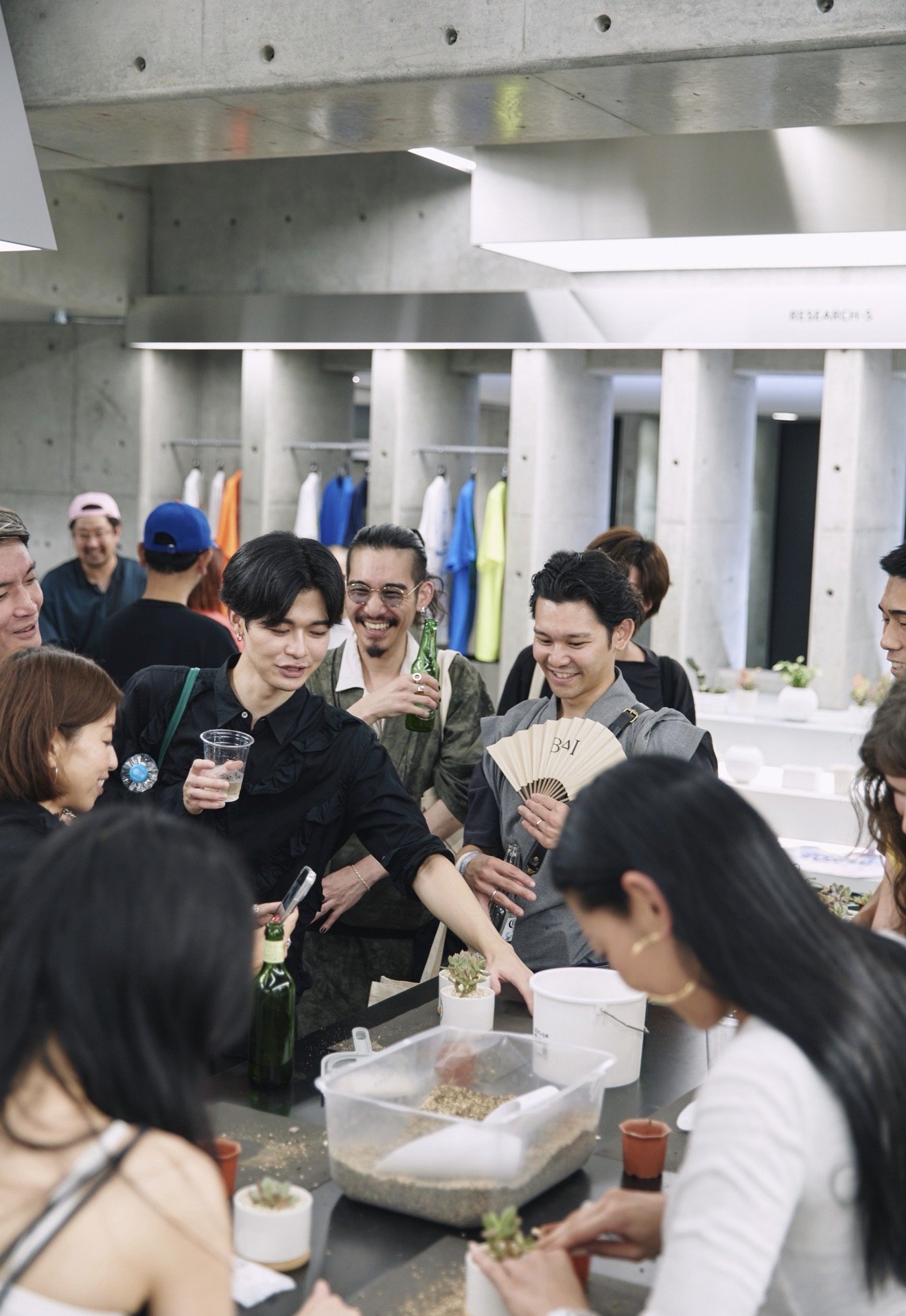 Group of people at a gathering, standing around a table with potted succulents, in a modern, industrial-style room with concrete walls and colorful clothing hanging in the background.