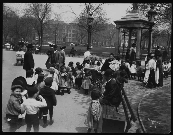 Children and adults gathering at a park in black and white, with some children in line for a carousel ride, and others playing or chatting. Trees and park structures are visible in the background.