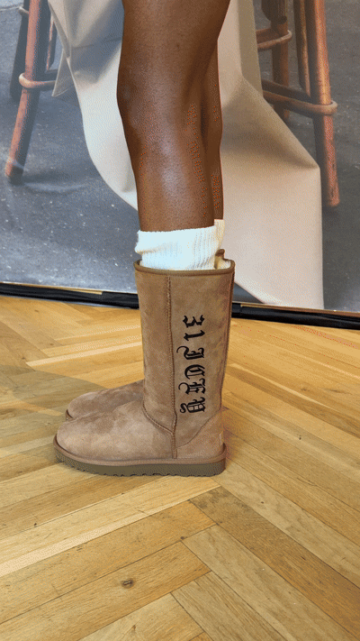 Close-up of a person wearing a beige winter boot with the words "BE LOYE" on the side, cream-colored sock, and a beige coat, standing on a wooden floor.
