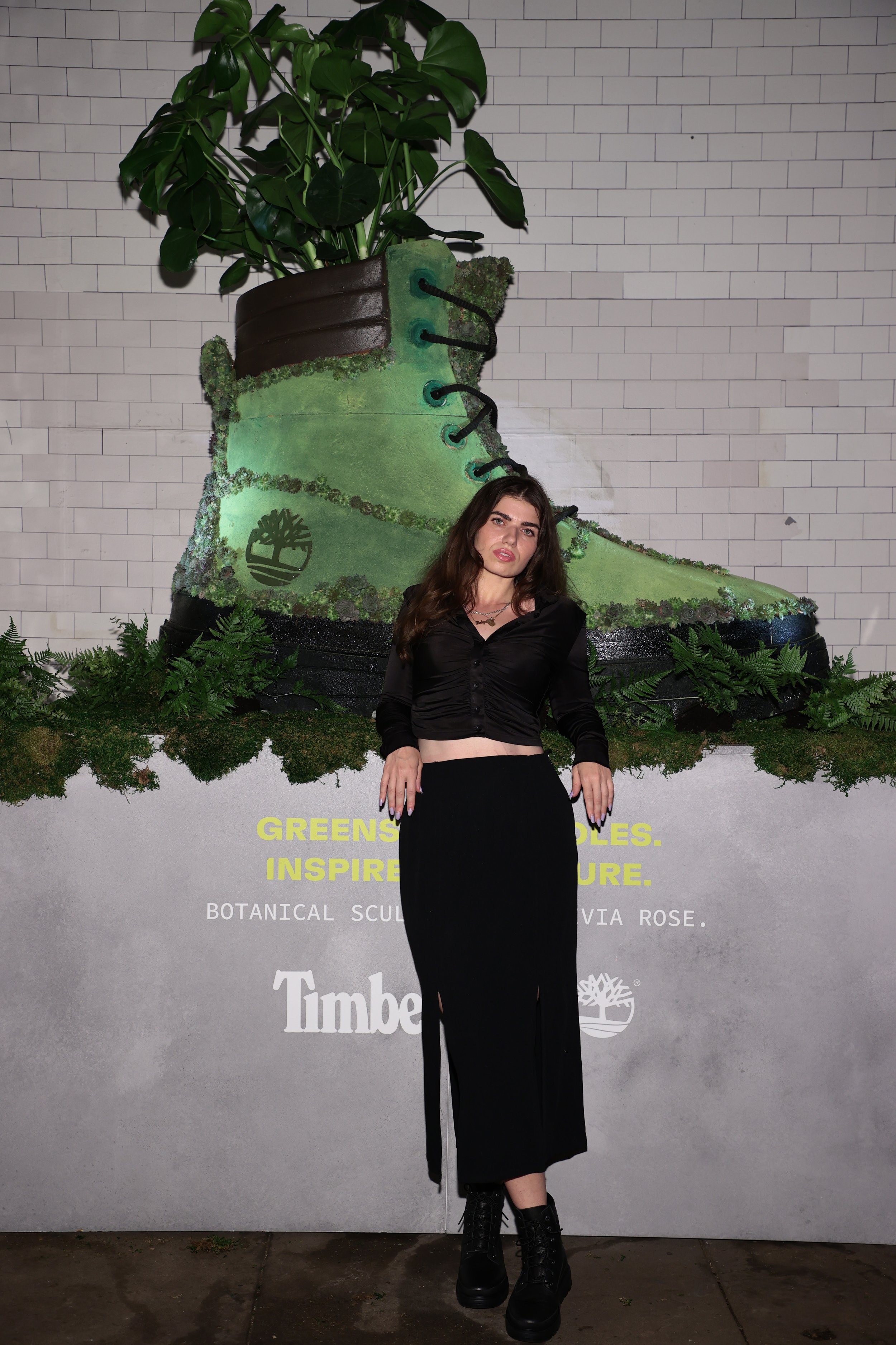A woman with long brown hair, wearing a black cropped blouse and black skirt, stands in front of a large decorative display featuring a giant green hiking boot with a plant growing out of the top. The background includes a white brick wall and a gray panel with yellow and white text, along with some green foliage at the base of the display.