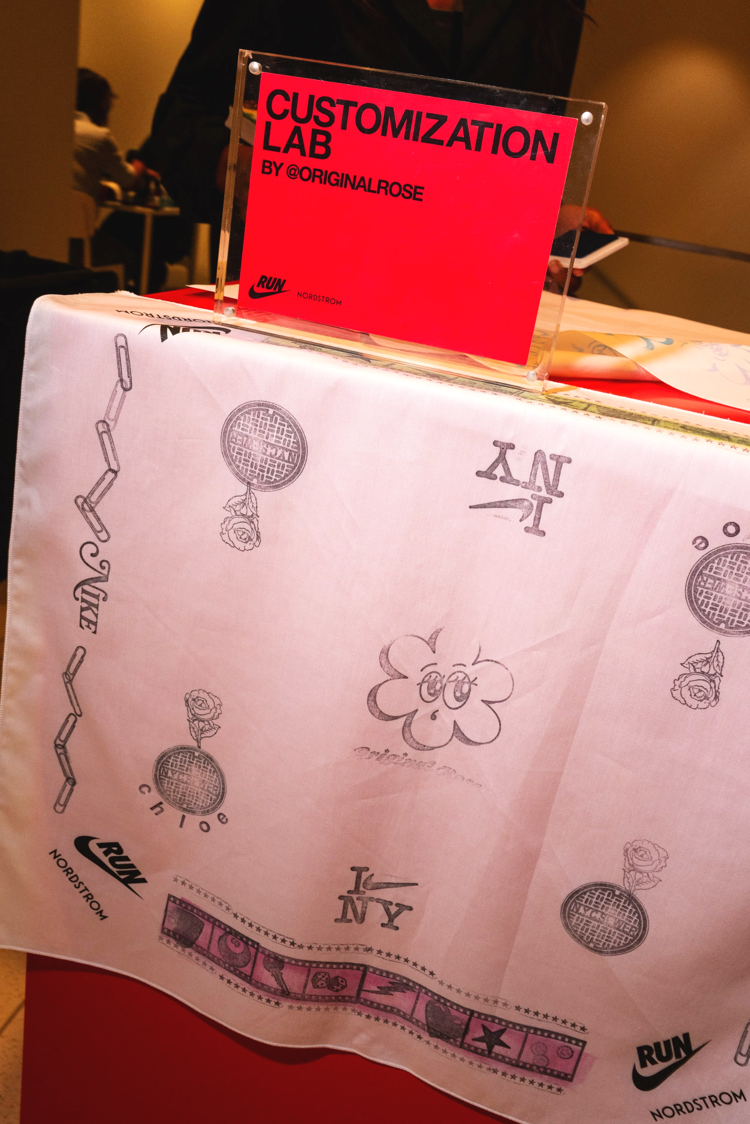 Sign that says 'Customization Lab by @OriginalRose' with Nike and Nordstrom logos, placed on a table with a pink and white tablecloth featuring sketches and patterns.