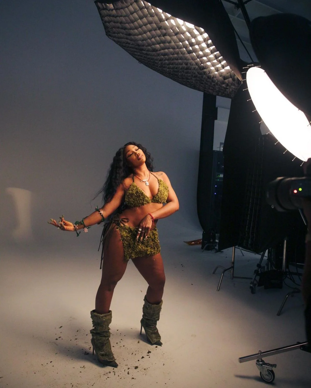 A woman poses in a photo studio wearing a costume made of moss with high heels, and her makeup and hair are styled elaborately. There is professional studio lighting equipment visible.