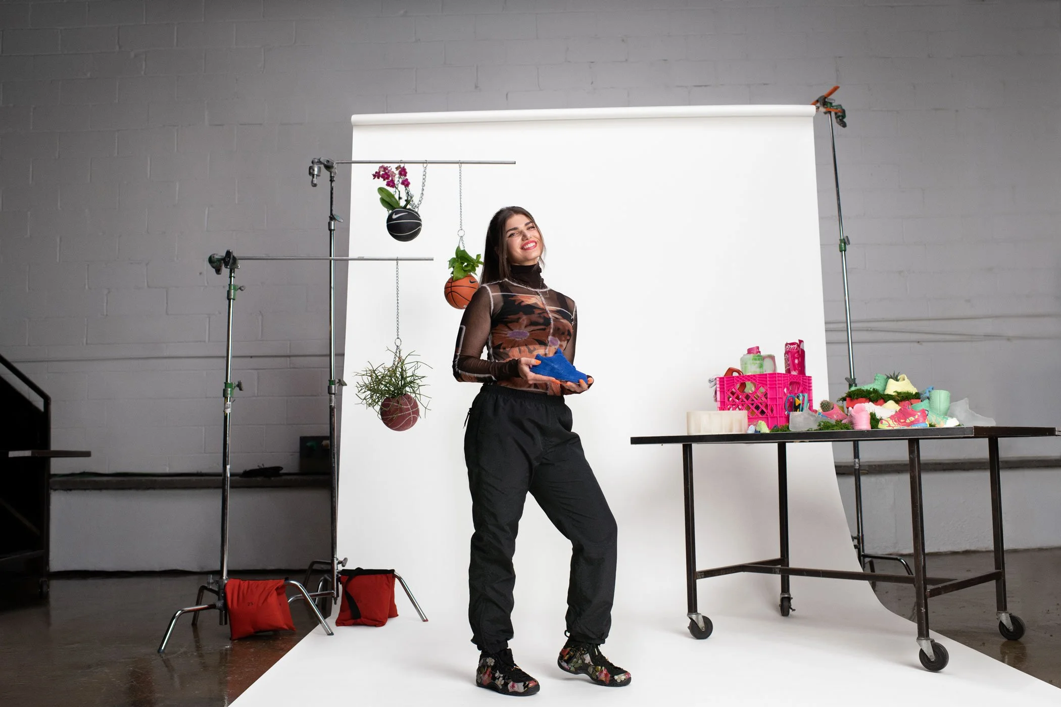 A woman standing in a photography studio with a white backdrop, holding a blue shoe, surrounded by plants, sports equipment, and various items on tables.