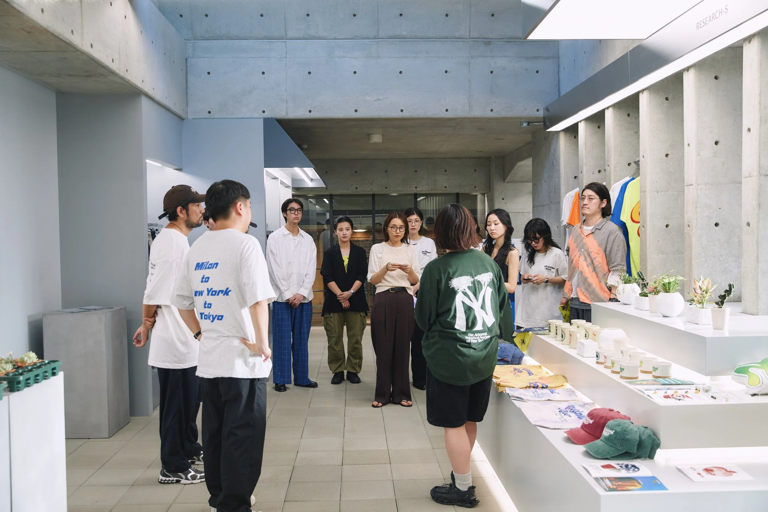 Group of young adults gathered around a display table inside a modern building, with concrete walls and plants, listening to a woman in a green shirt speaking.
