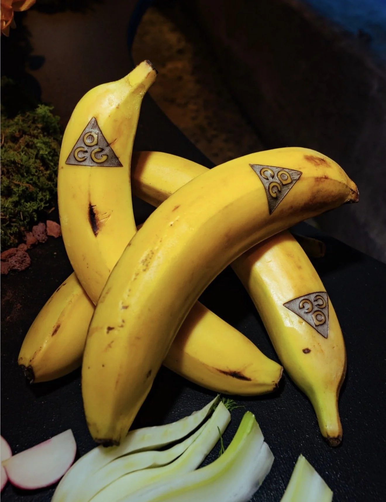 A bunch of yellow bananas with the AAA logo on them, placed on a black surface with some sliced onions nearby.