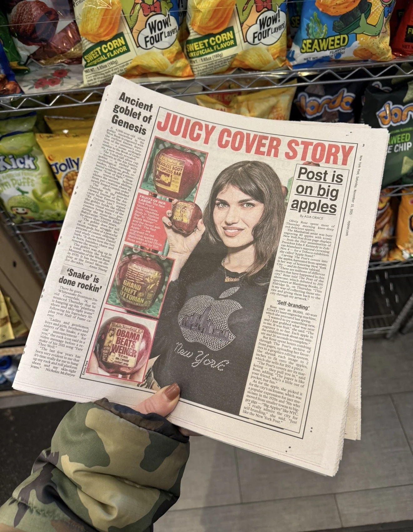 Person holding a newspaper with a woman on the cover and headlines about apples and snacks, with snack bags on store shelves in the background.