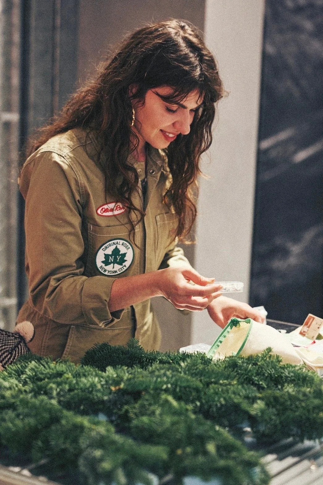 A woman with long dark curly hair, wearing a tan military-style jacket with patches, is smiling and looking down at a table with greenery and wrapped gifts or packages, inside a building.