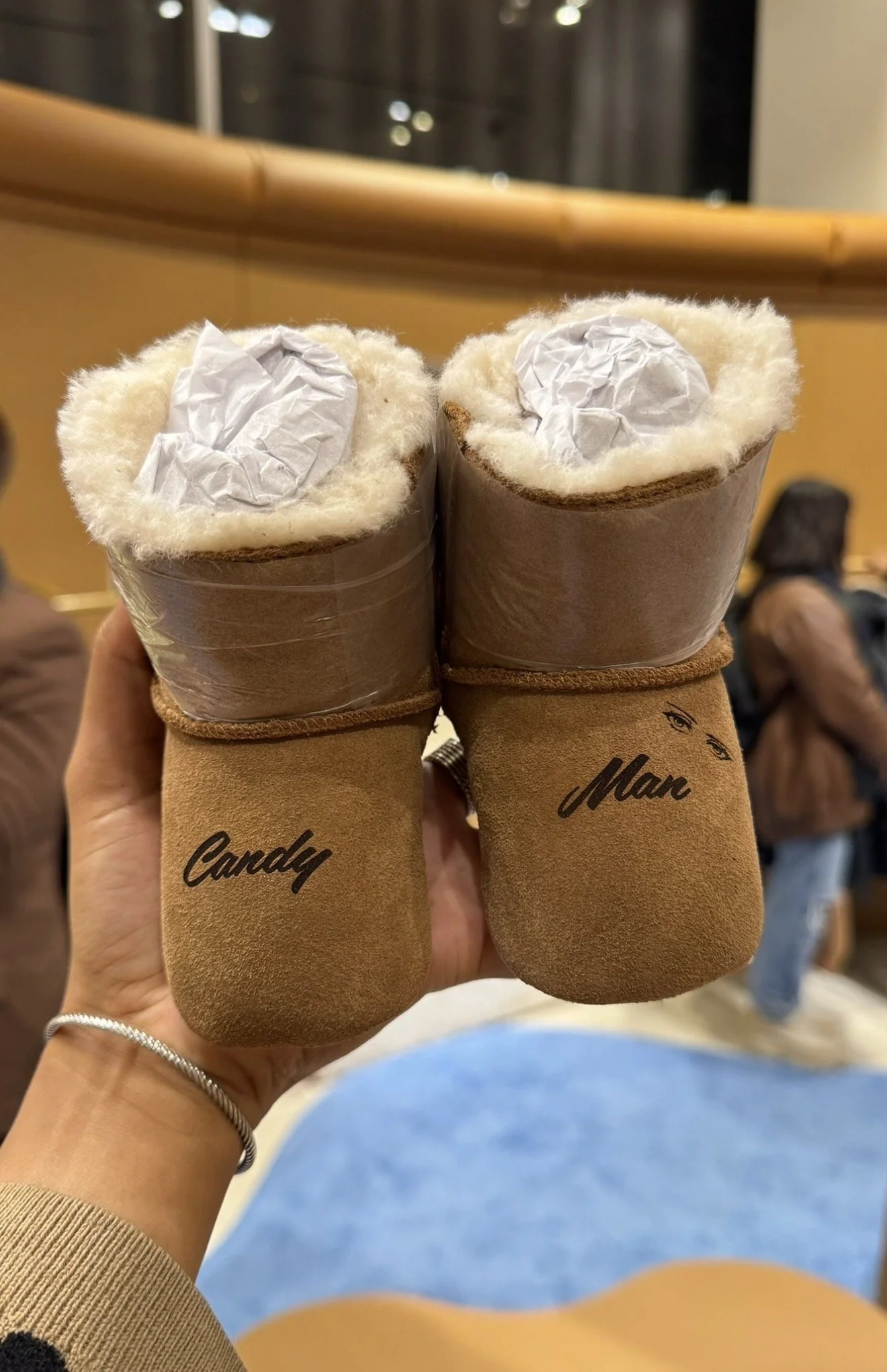 Pair of cozy winter boots with fluffy lining and paper insert, labeled 'Candy' and 'Man', held in someone's hand inside a waiting area with people sitting in the background.