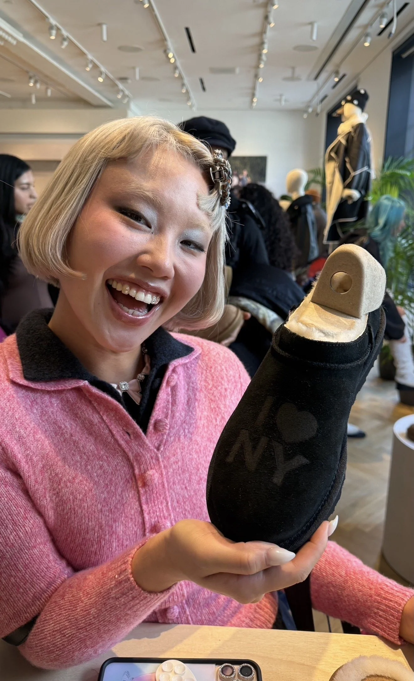 A woman with a short blonde bob haircut, smiling and holding a black shoe with a beige sole, inside a retail store or gallery with mannequins and other customers in the background.