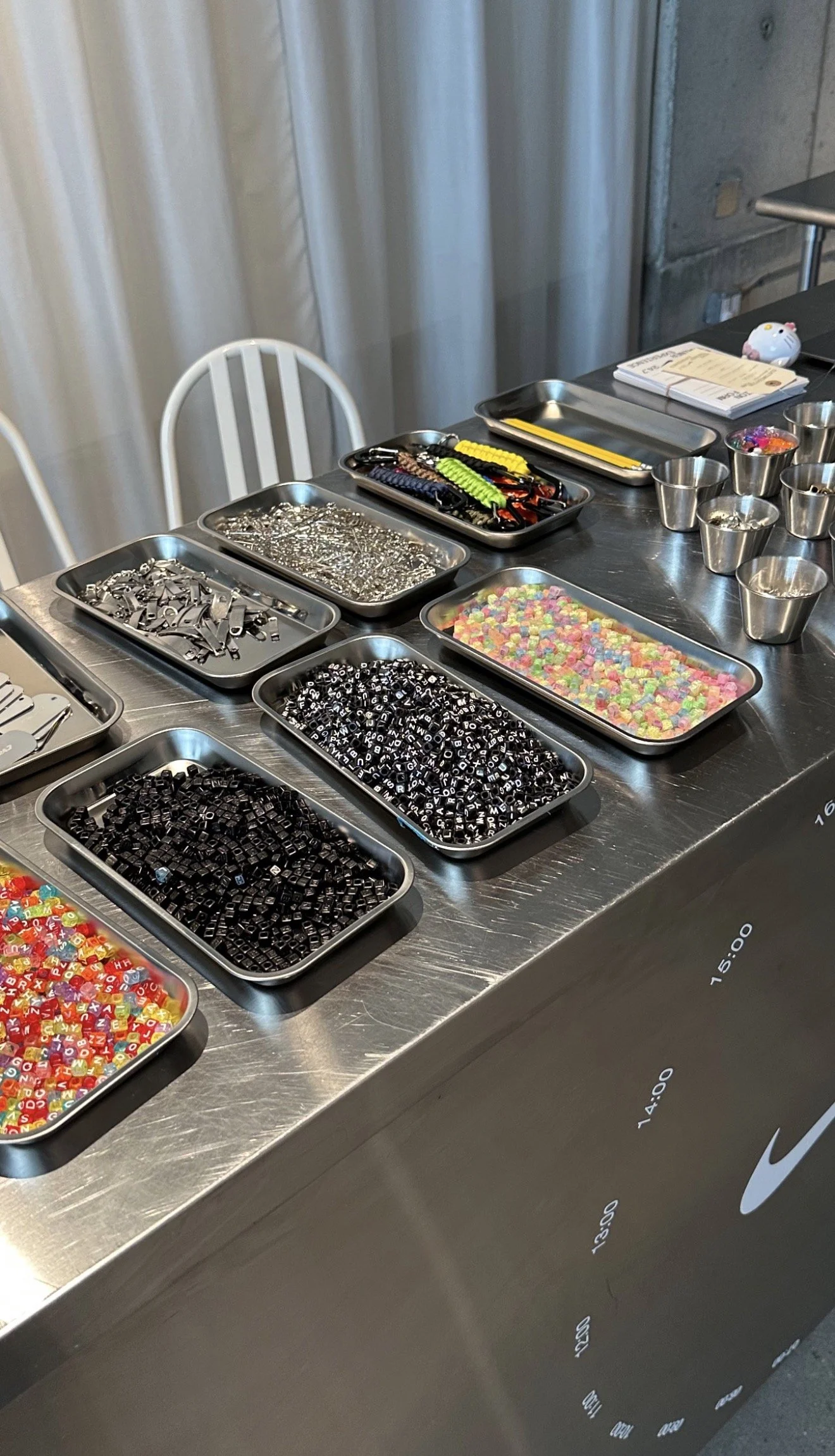 A stainless steel table with trays of various alphabet beads, small colorful beads, and jewelry making supplies in a room with white chairs and a concrete wall behind.