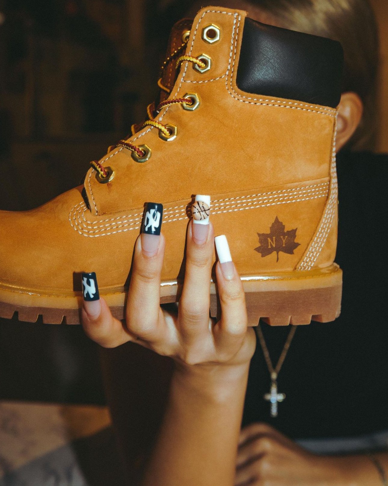 A hand with decorative nail art holding a tan-colored hiking boot with black ankle collar, gold eyelets, and a NY maple leaf logo, against a dark background.