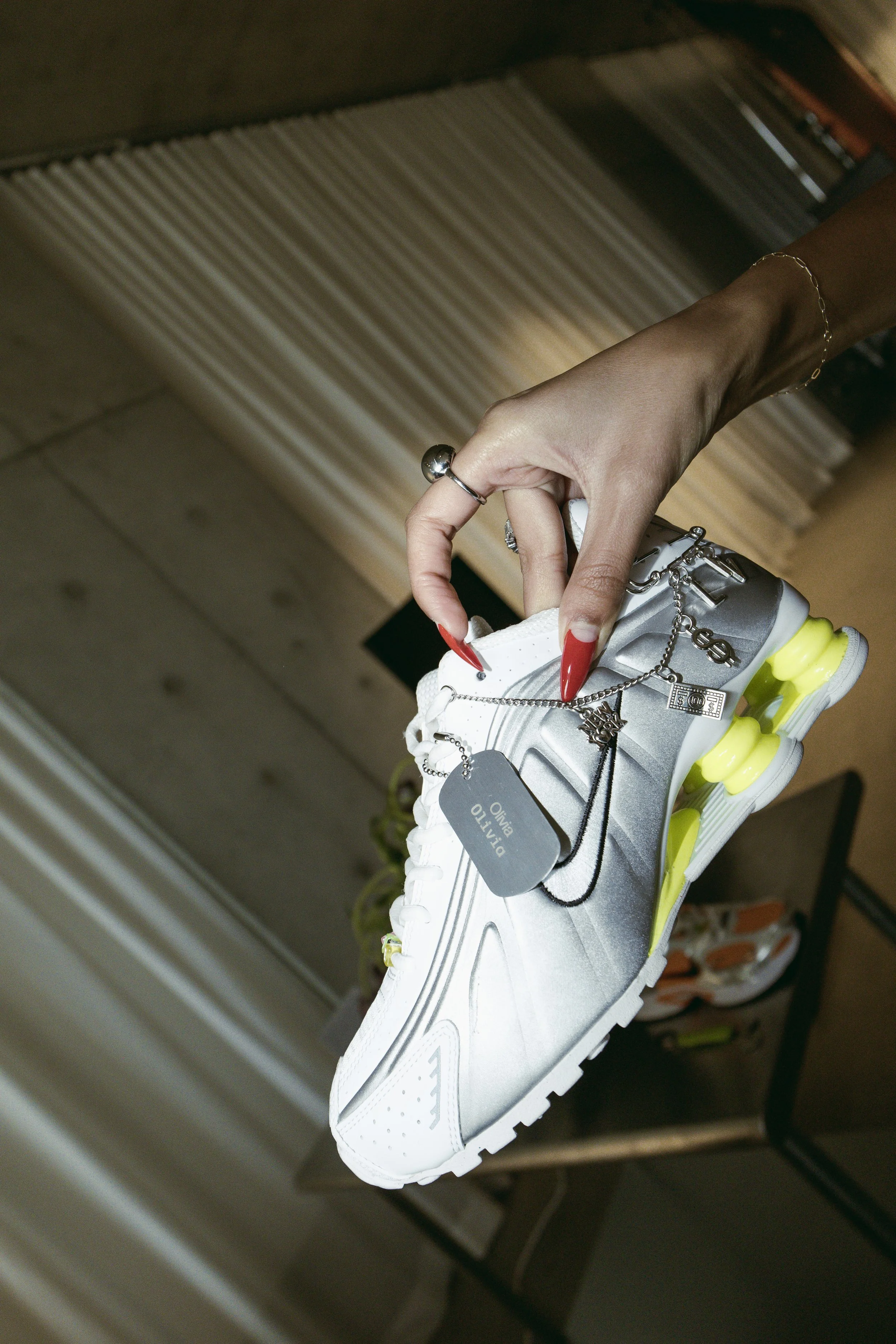 A person's hand with red nails and jewelry holding a metallic silver and white athletic shoe with yellow accents, containing a tag that says 'DILITO OLIVIA' and decorative chains attached to the laces.