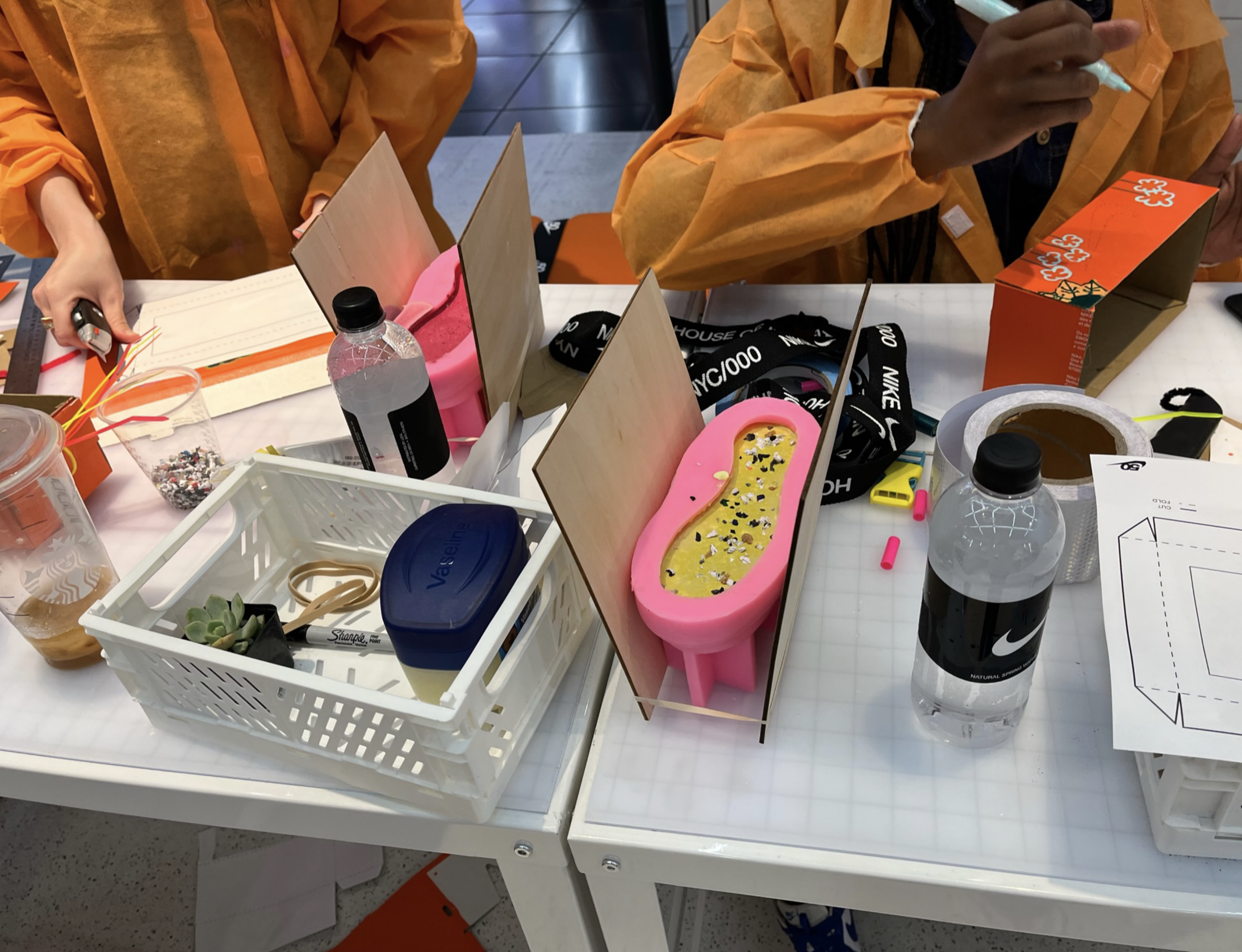 A workspace with various craft supplies, including bottles of water, a container of honey, a small container of sprinkles, a pink and yellow salt or sugar holder, scissors, markers, tape, a piece of paper, and a small succulent plant. Several people 