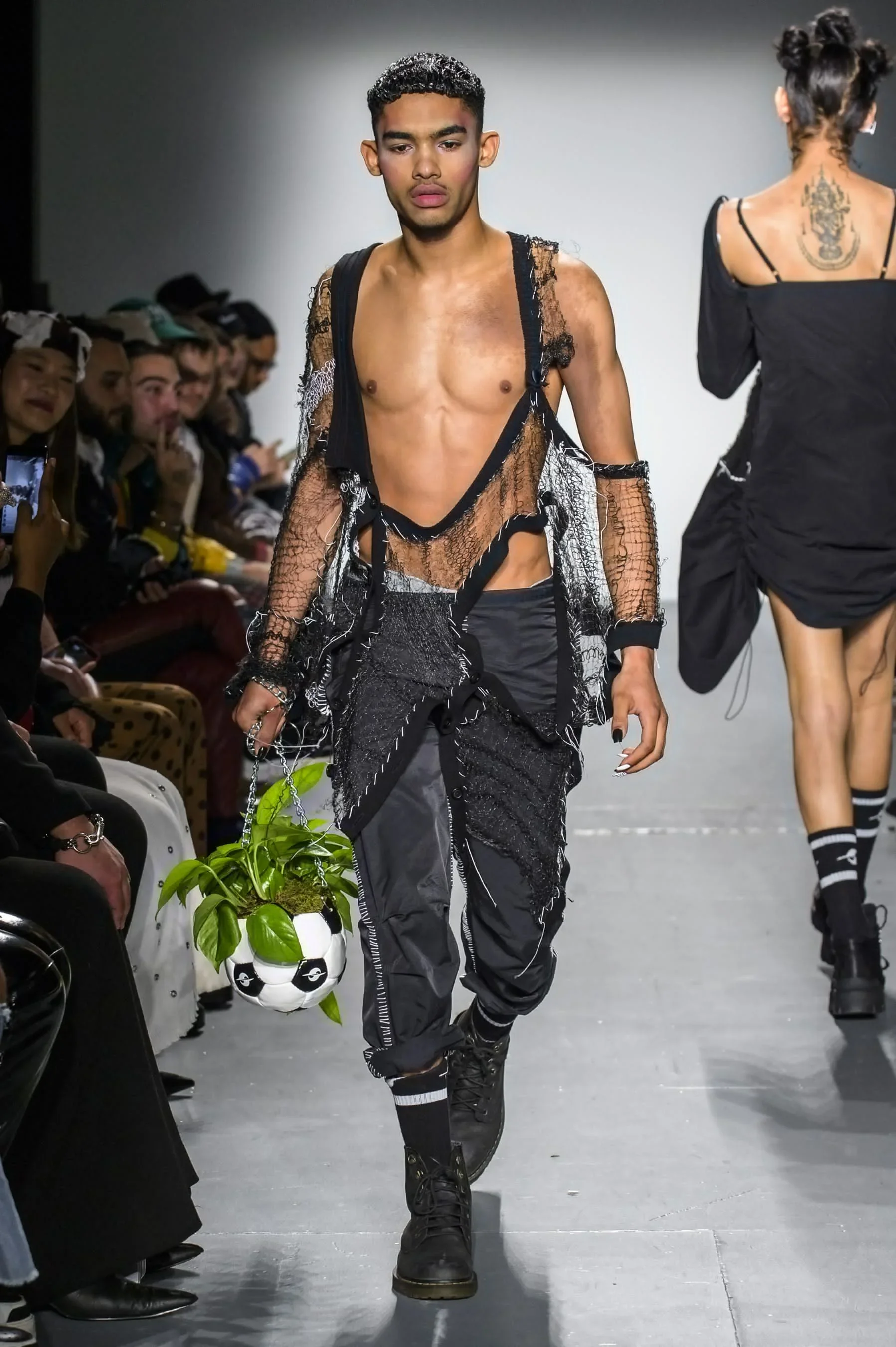 Male model walking down a runway during a fashion show, holding a small plant in a soccer ball pot, wearing distressed black clothing and black boots, with an audience seated on both sides.