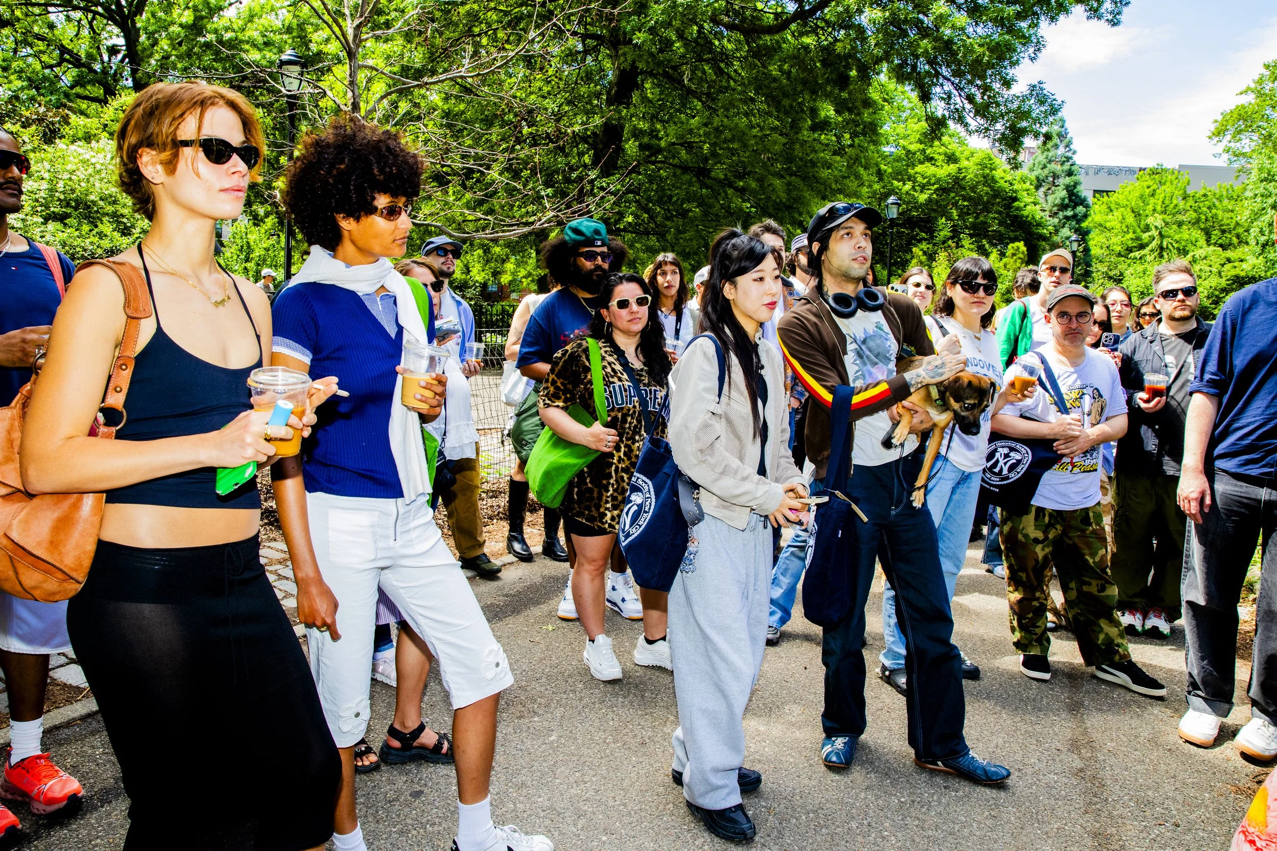 A diverse group of people attending an outdoor event in a park, some holding drinks, with lush green trees in the background.